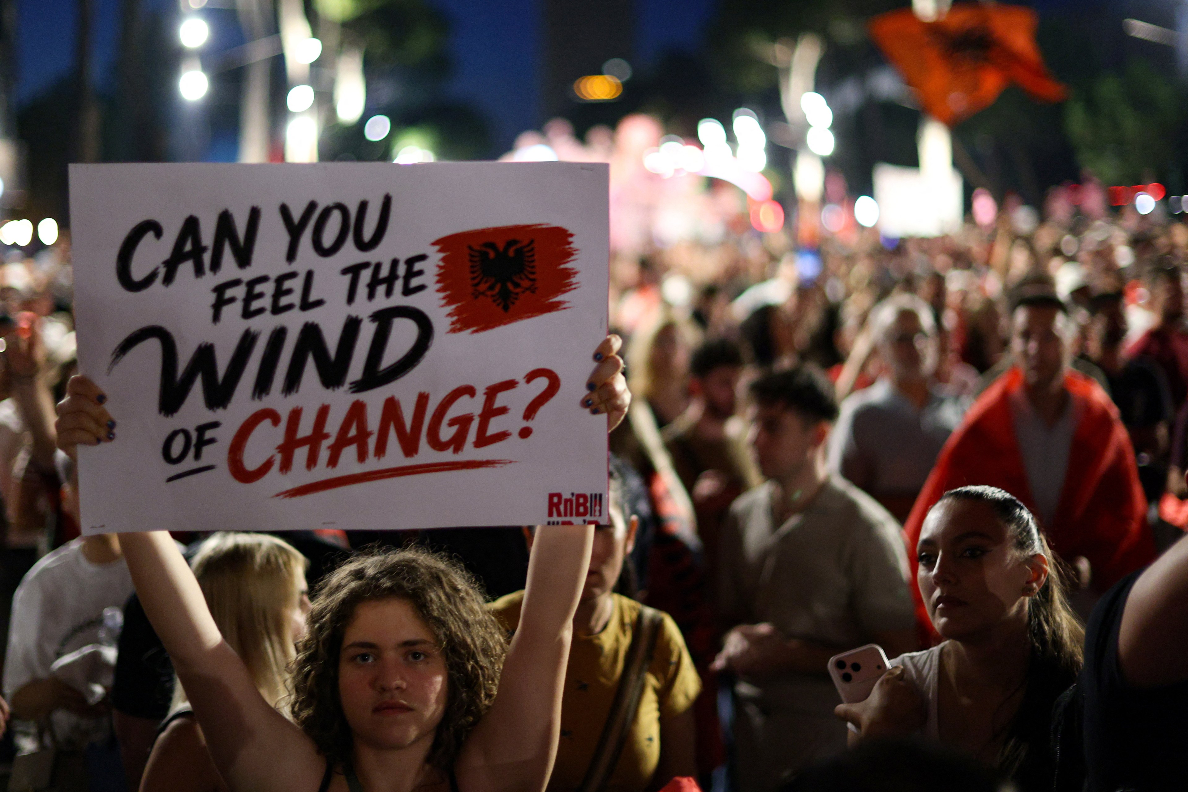 Young person holding a sign reading 'Can you feel the wind of change?' with the Albanian flag, surrounded by a large crowd at a nighttime protest in Tirana, Albania.