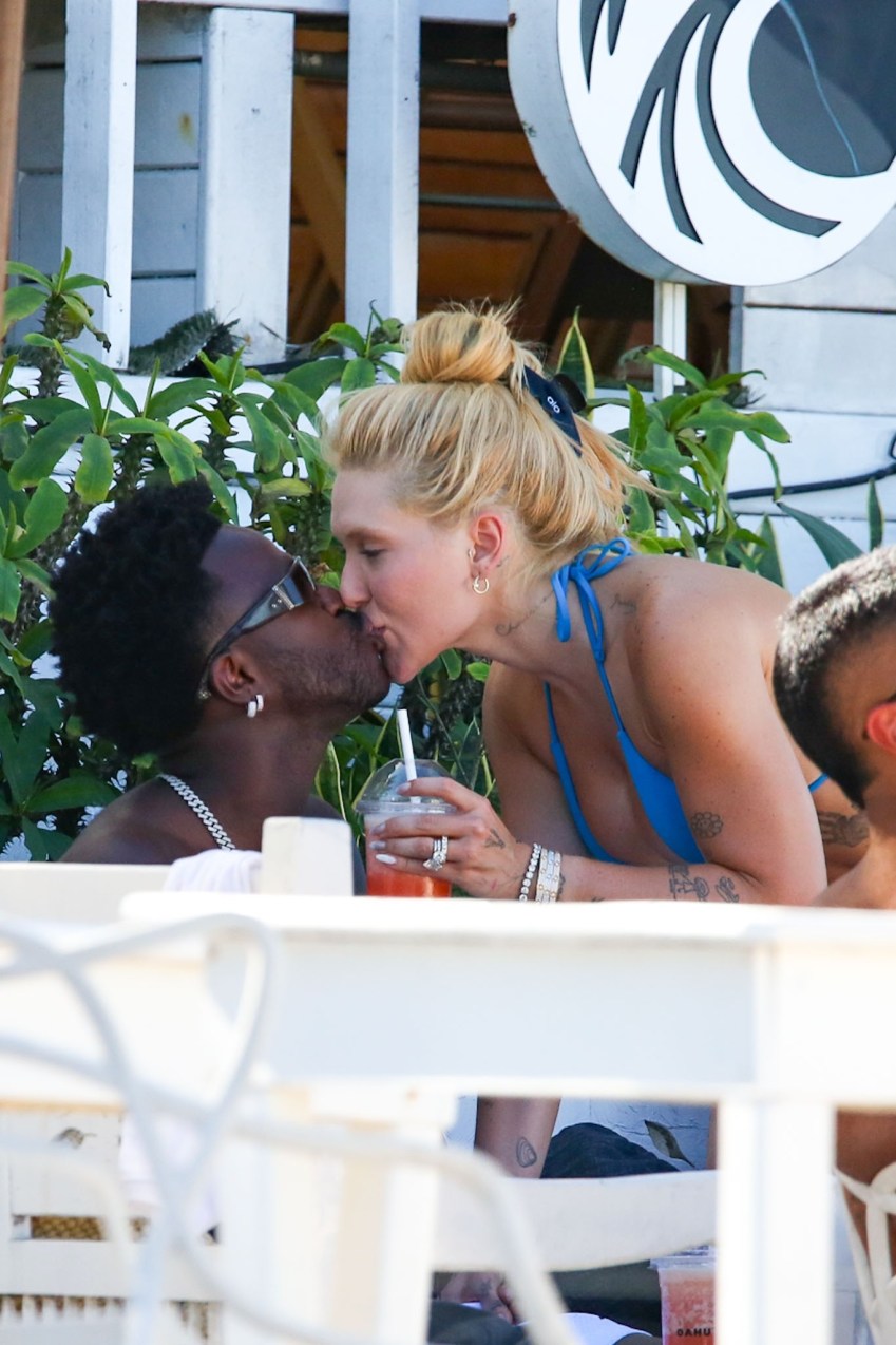A man and a woman in a blue bikini top share a kiss at an outdoor beach venue, she holds an iced drink.