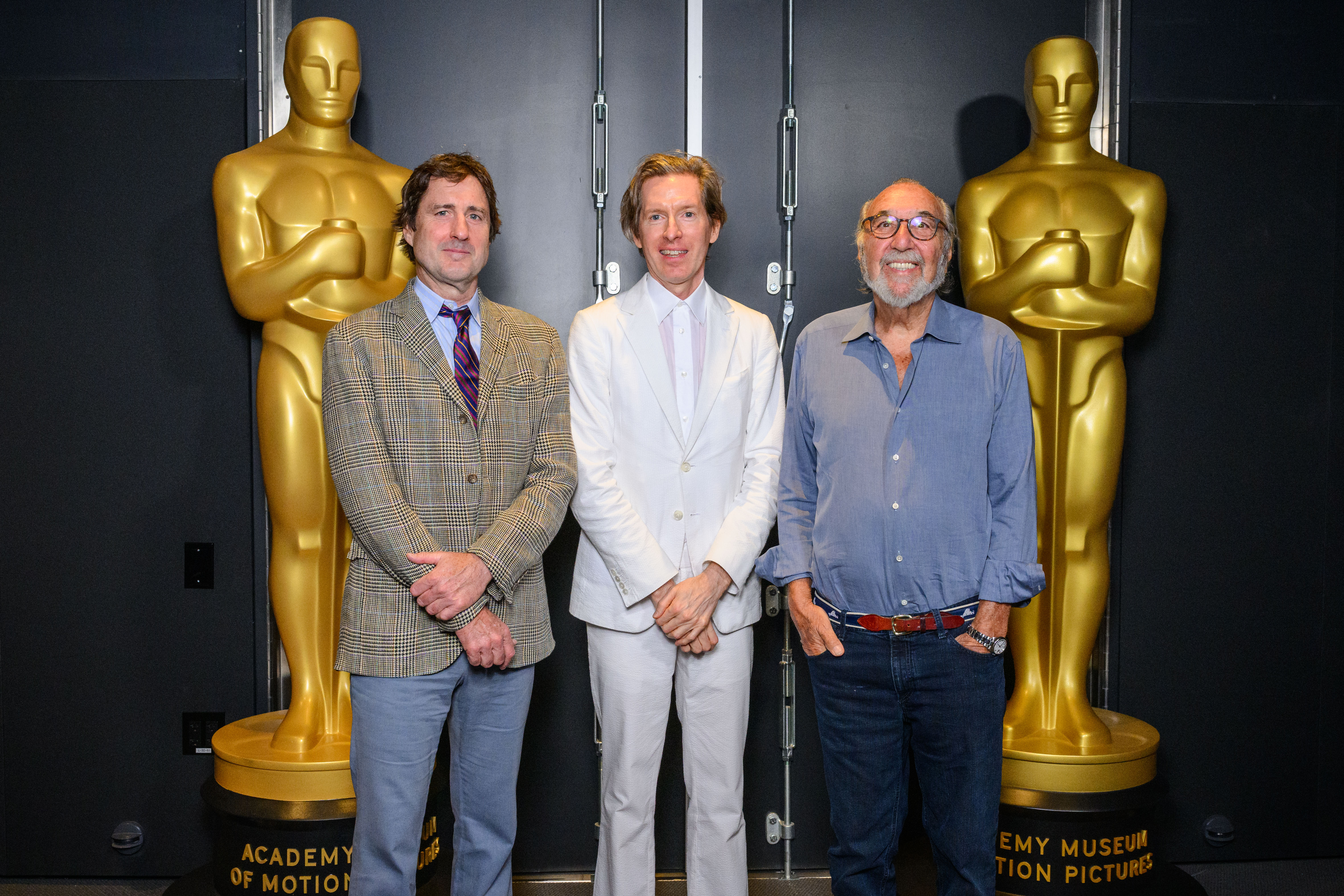 Three men stand smiling between two large golden Oscar statues at the Academy Museum of Motion Pictures.