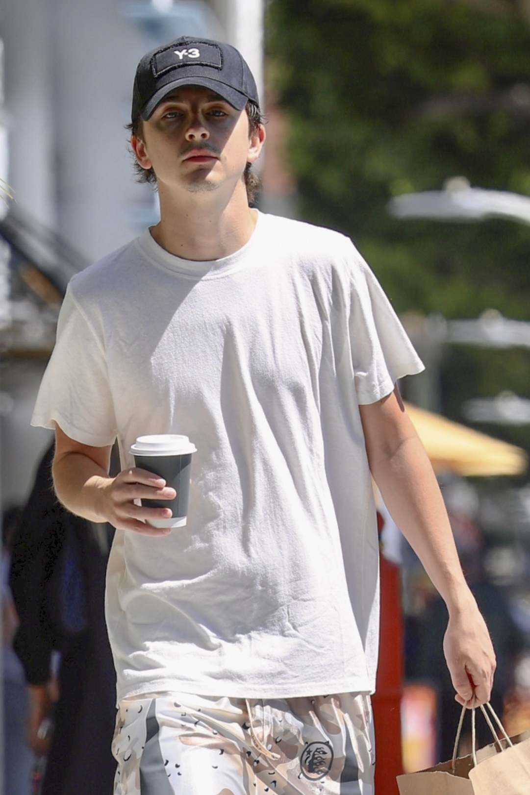 Timothée Chalamet in a white T-shirt, black cap, and white sneakers holding a coffee cup and paper bag outside a café in Beverly Hills