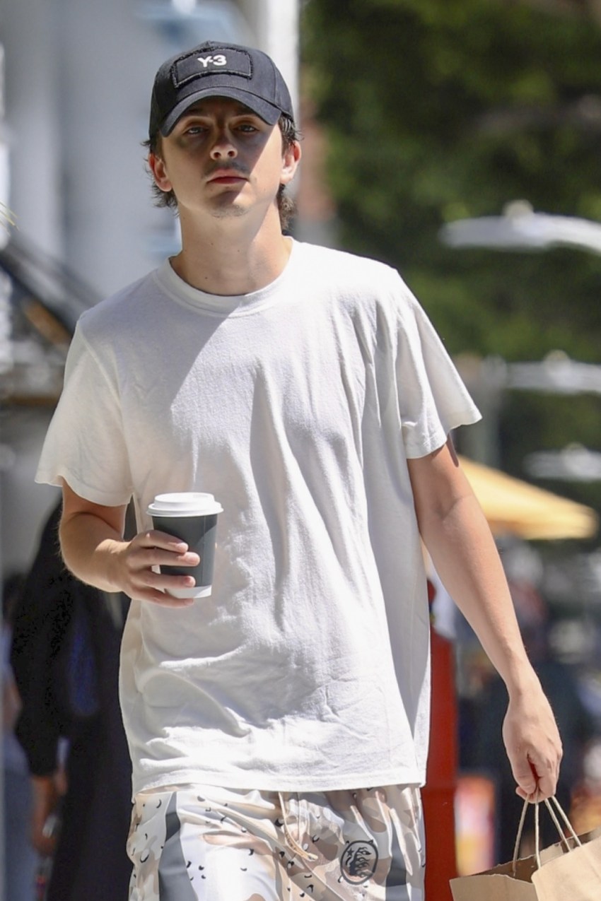 Timothée Chalamet in a white T-shirt, black cap, and white sneakers holding a coffee cup and paper bag outside a café in Beverly Hills