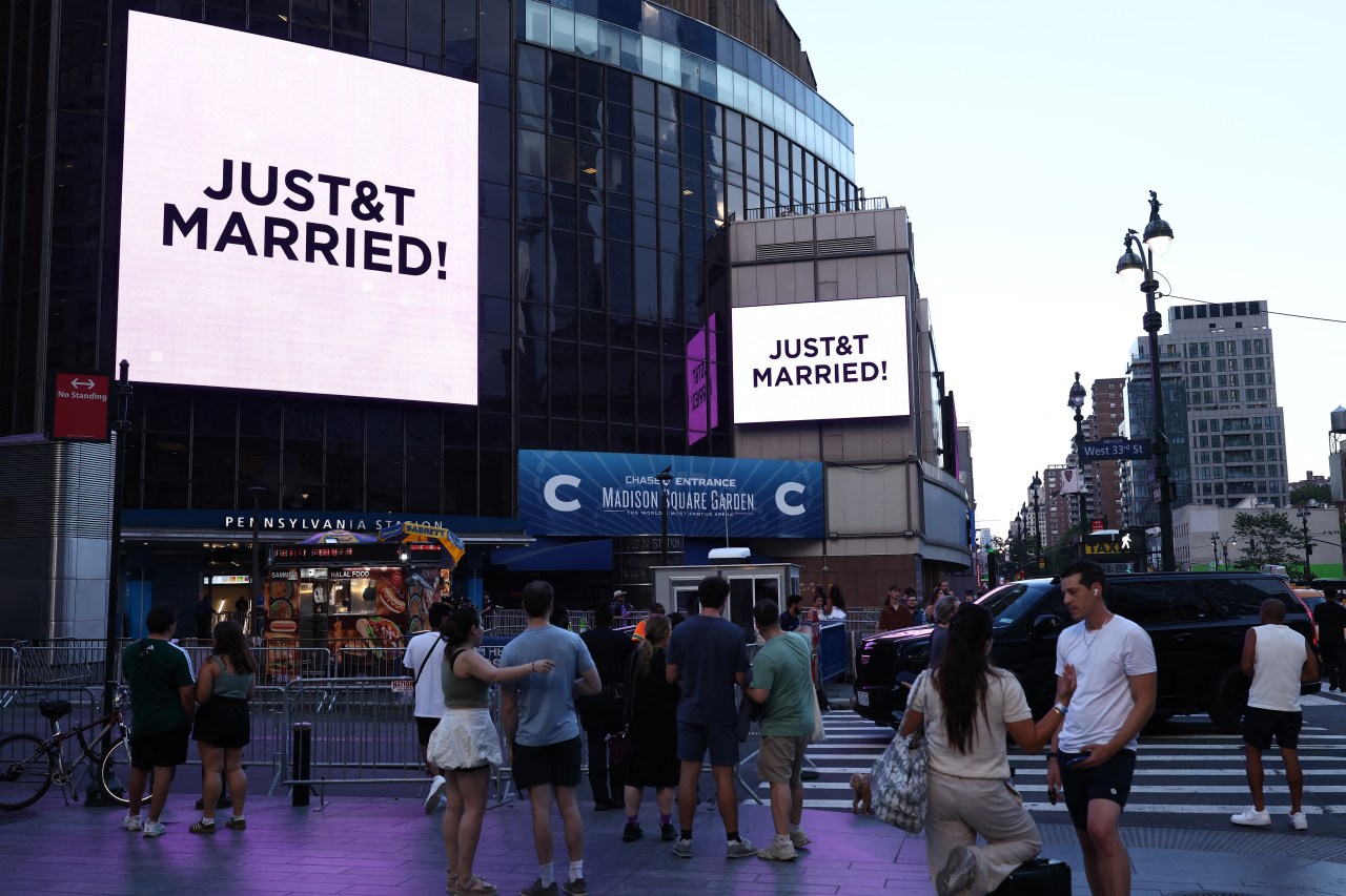 Crowd outside Madison Square Garden watching large screens displaying "JUST&T MARRIED!" celebrating Taylor Swift and Travis Kelce's wedding