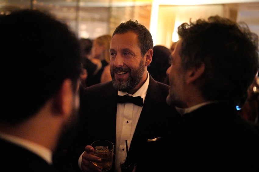 Bearded man in a tuxedo with bow tie laughing and holding a whiskey glass at a crowded upscale party.