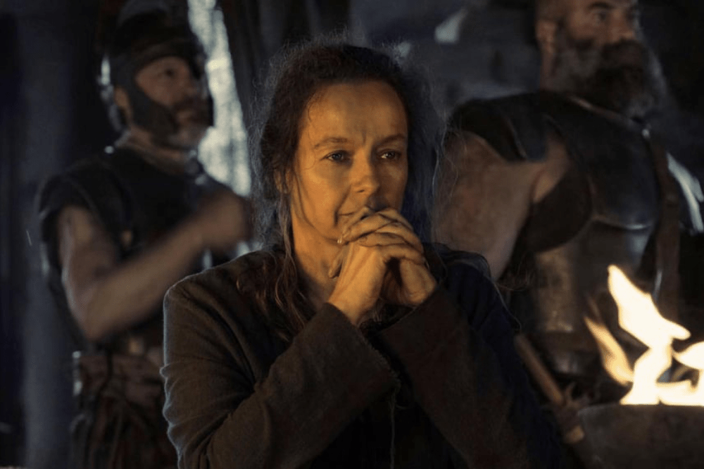 A worried woman in dark robes holds clasped hands to her mouth, with armored men blurred in the background and firelight to her right.