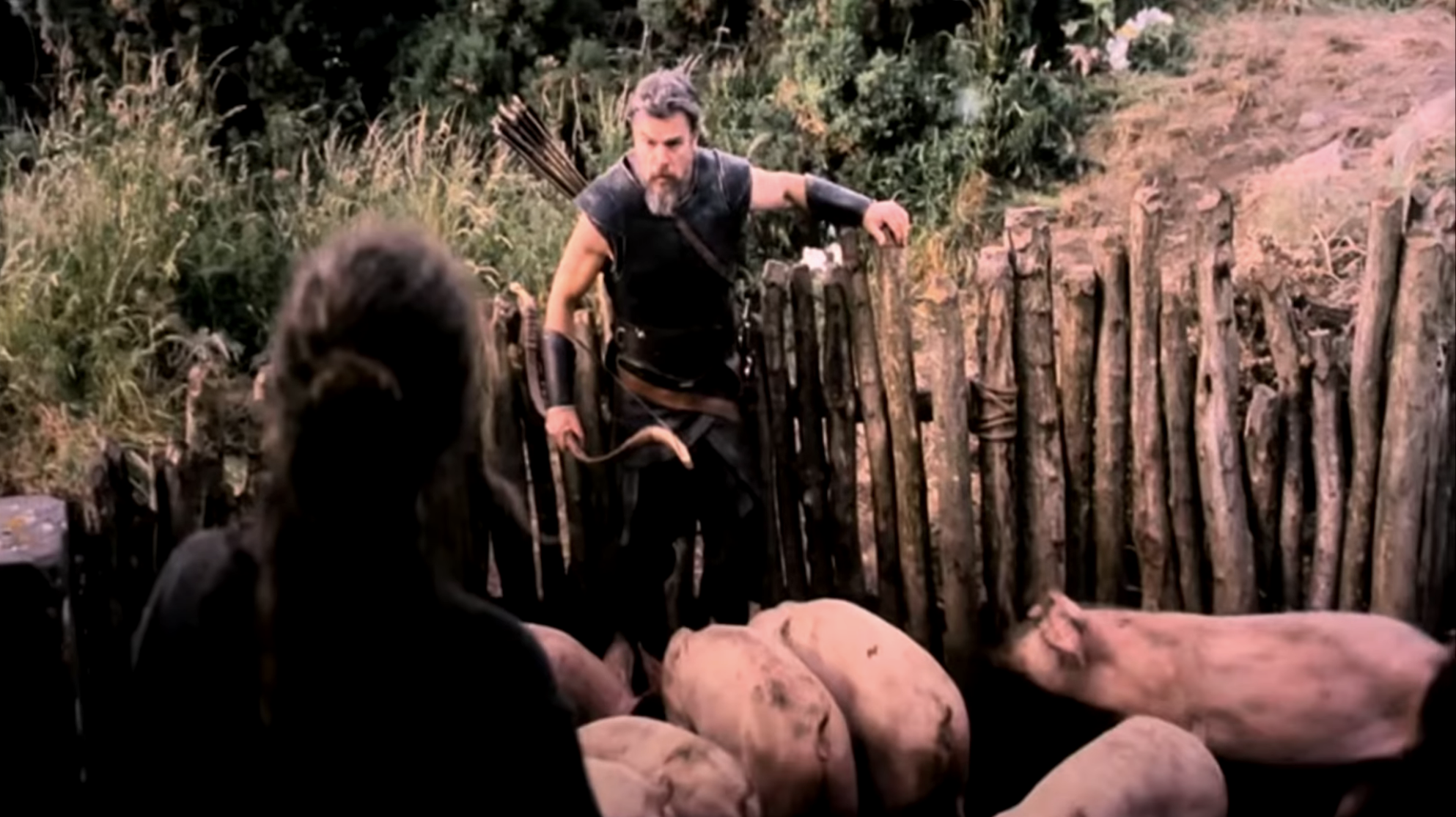 A bearded man armed with a bow and quiver leans over a rough wooden fence, facing a dark-haired figure, with bodies lying in the foreground.