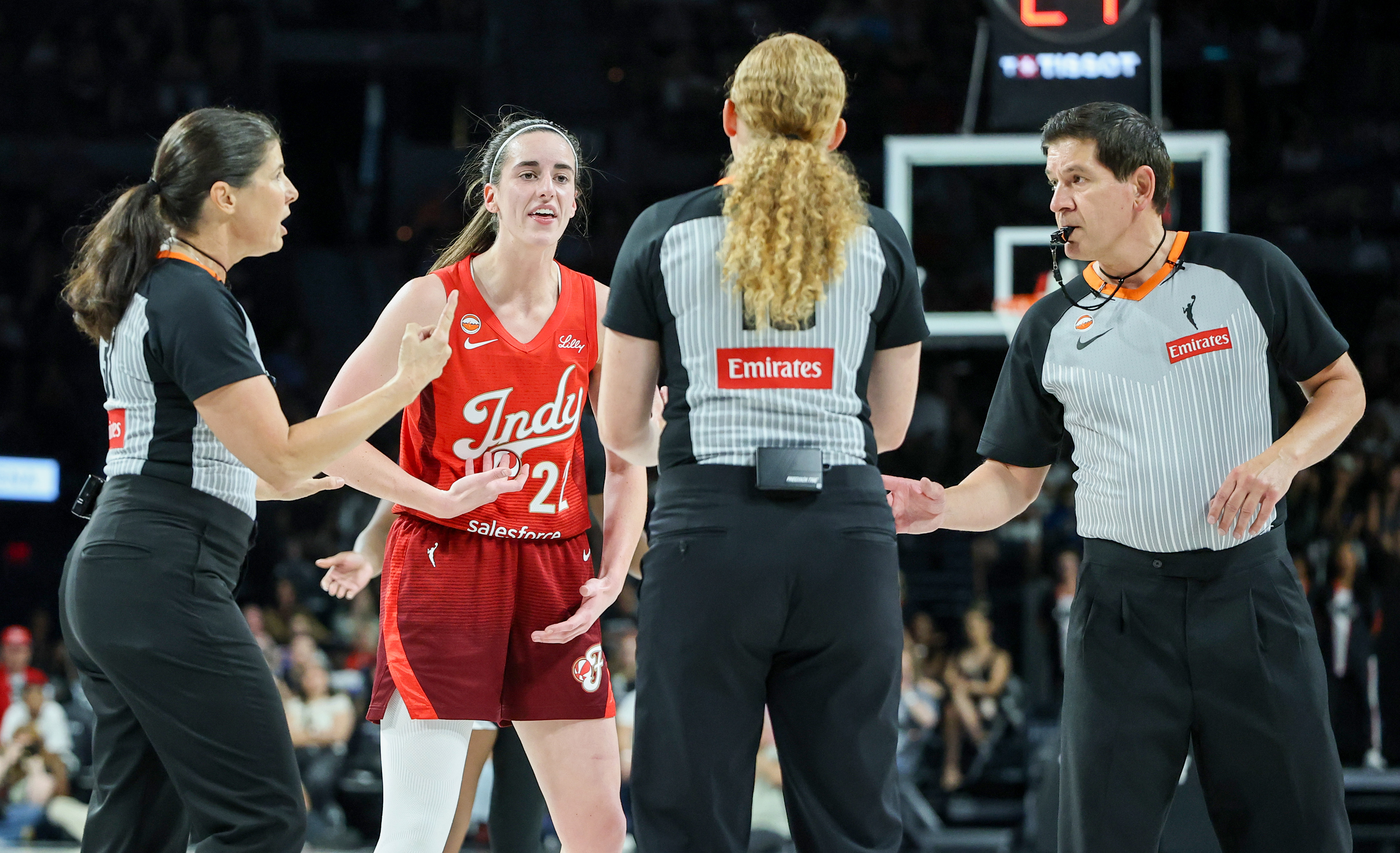 Caitlin Clark in a red Indy jersey gestures and argues with three referees on a WNBA court.