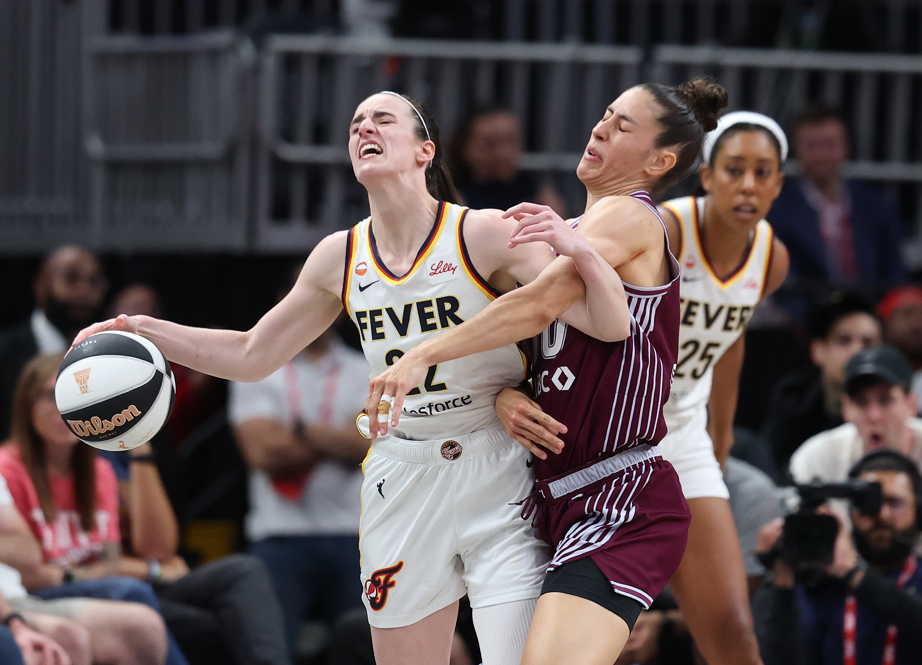 Caitlin Clark in a white Fever jersey holds the ball while being fouled by an opposing player in a maroon uniform.