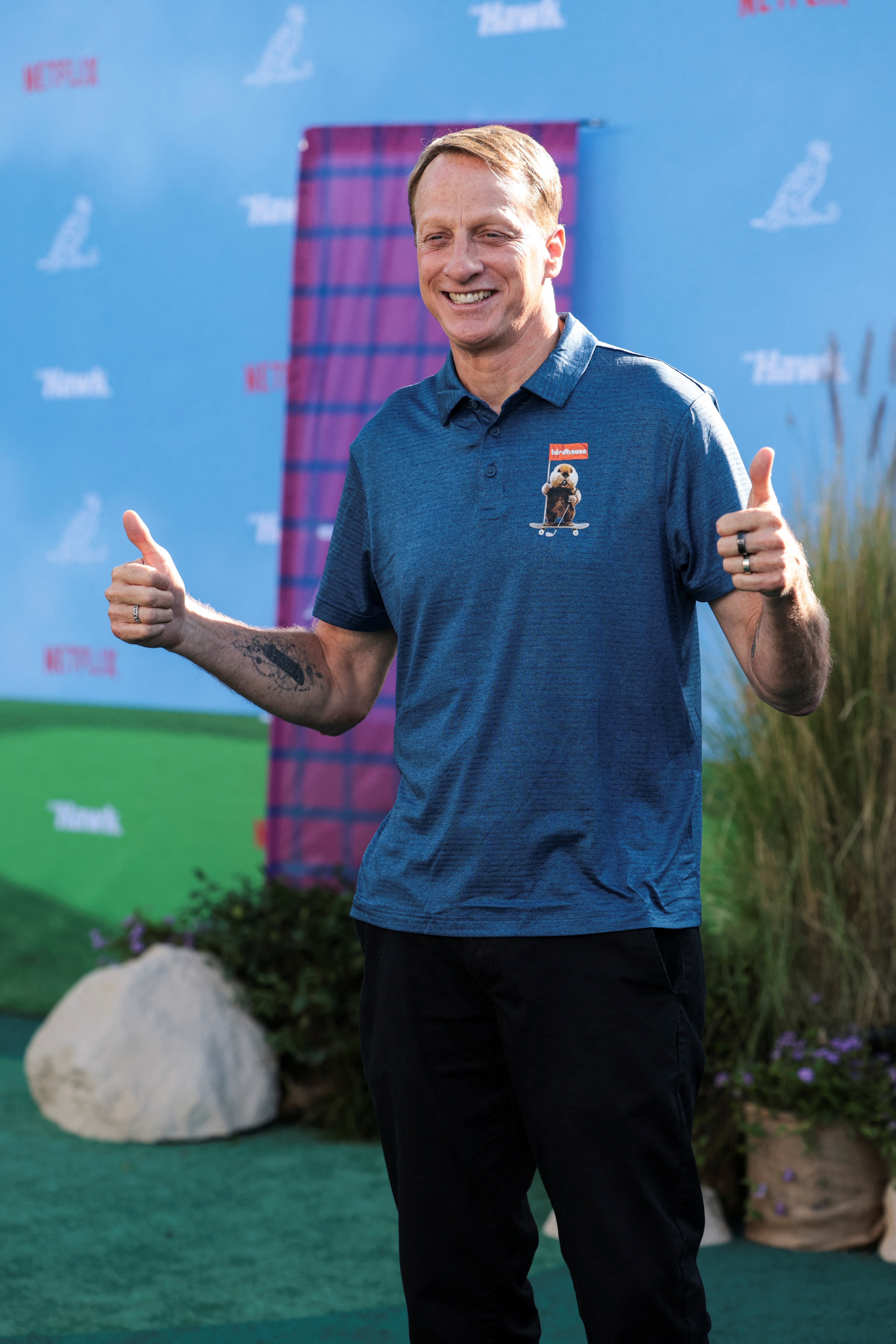 Tony Hawk smiling and giving two thumbs up at a Netflix premiere event, wearing a blue polo shirt with a Birdhouse logo.