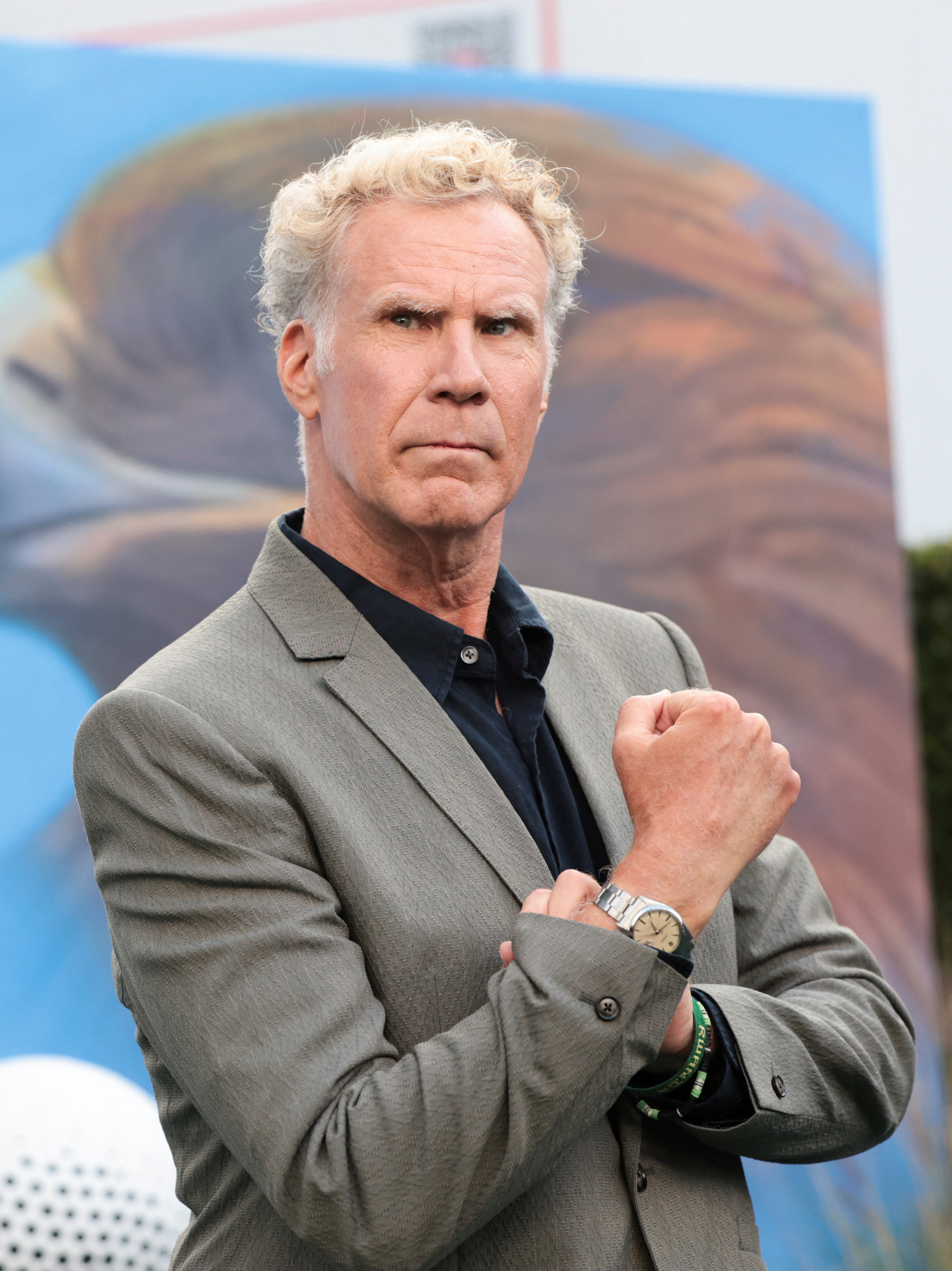 Will Ferrell in a grey blazer and black shirt, arms crossed, at a premiere event with a large bird mural behind him.