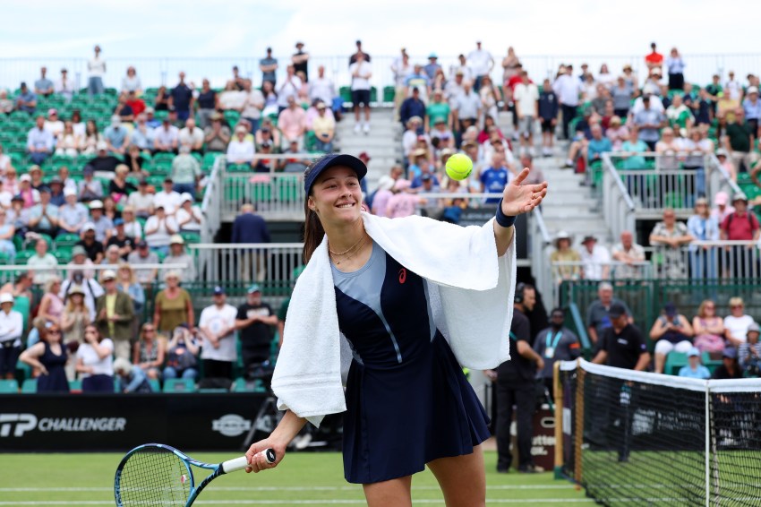 Female tennis player in navy Asics dress and cap smiling as she tosses a tennis ball into a packed crowd on an outdoor grass court.