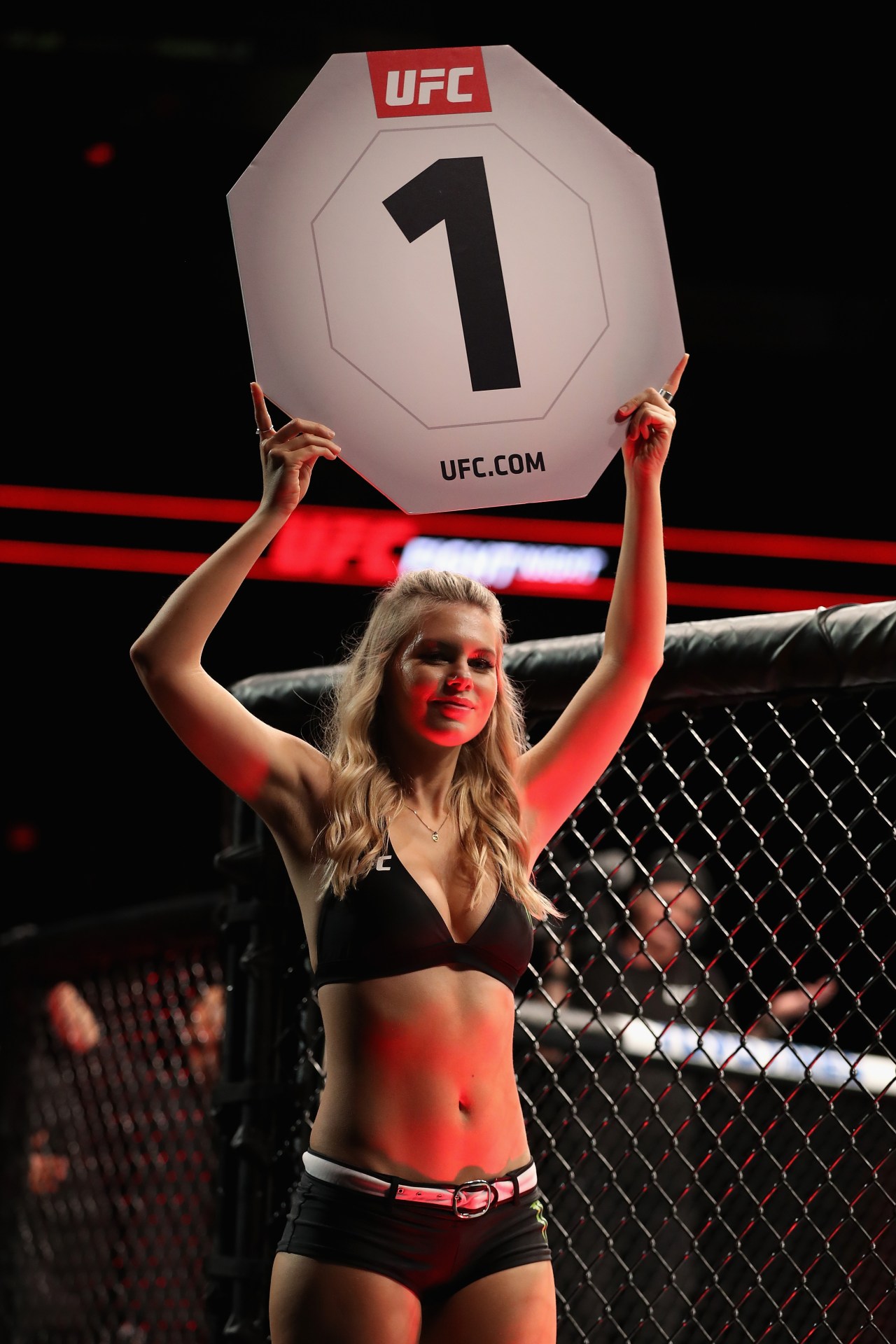 A blonde UFC Octagon Girl in a black bikini top and shorts holds a round 1 sign above her head inside the UFC cage.