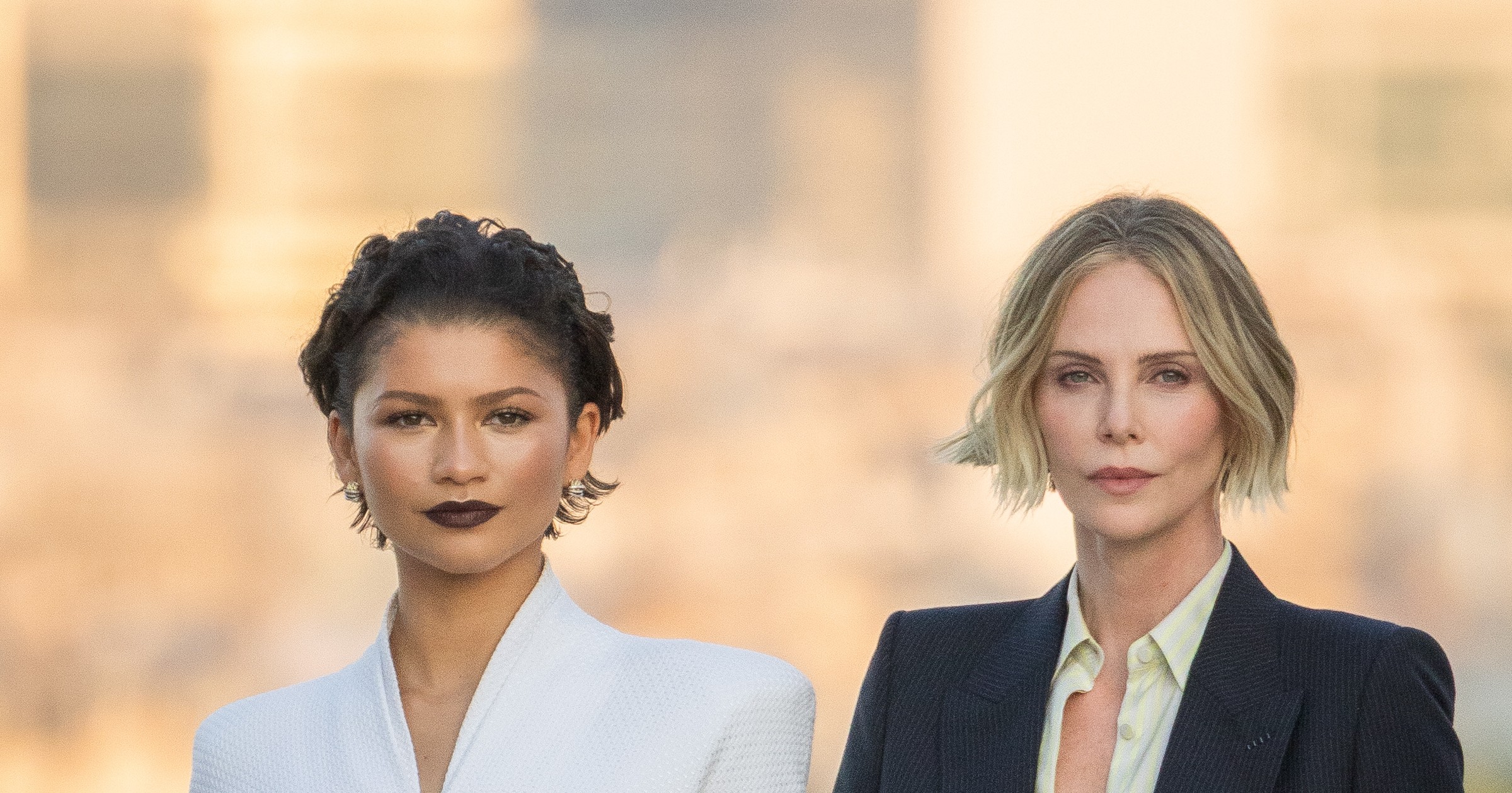 Zendaya in a white long-sleeve dress and Charlize Theron in a navy blazer and light blue satin shorts standing together outdoors at the Trocadero in Paris.