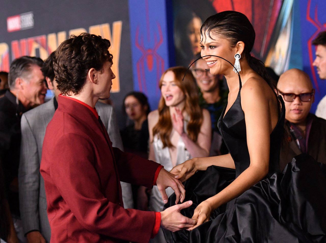 Zendaya in a black satin gown holds hands and laughs with a man in a red suit at a Marvel Spider-Man premiere, with a crowd visible behind them.