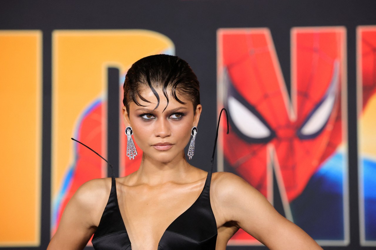 Woman in black satin dress with slicked-back hair and long silver drop earrings posing in front of a Spider-Man movie premiere backdrop.