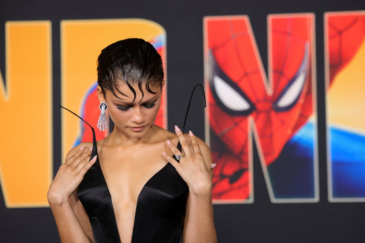 Zendaya in a black satin deep-V corset holds its straps, looking down, in front of a Spider-Man: Brand New Day premiere backdrop.