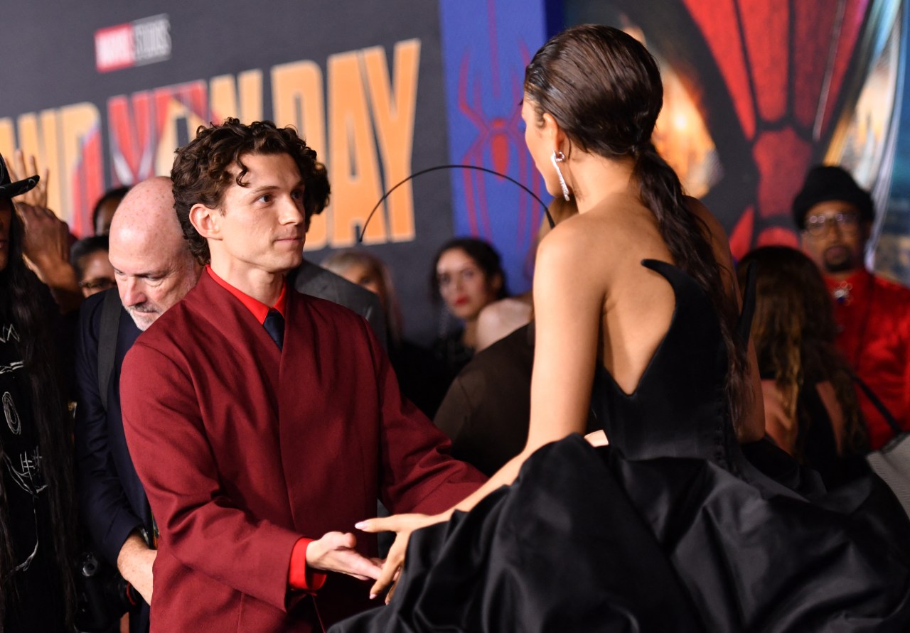 Tom Holland in a red jacket holds hands with Zendaya in a black gown at a Marvel Studios premiere, Spider-Man signage visible behind them.