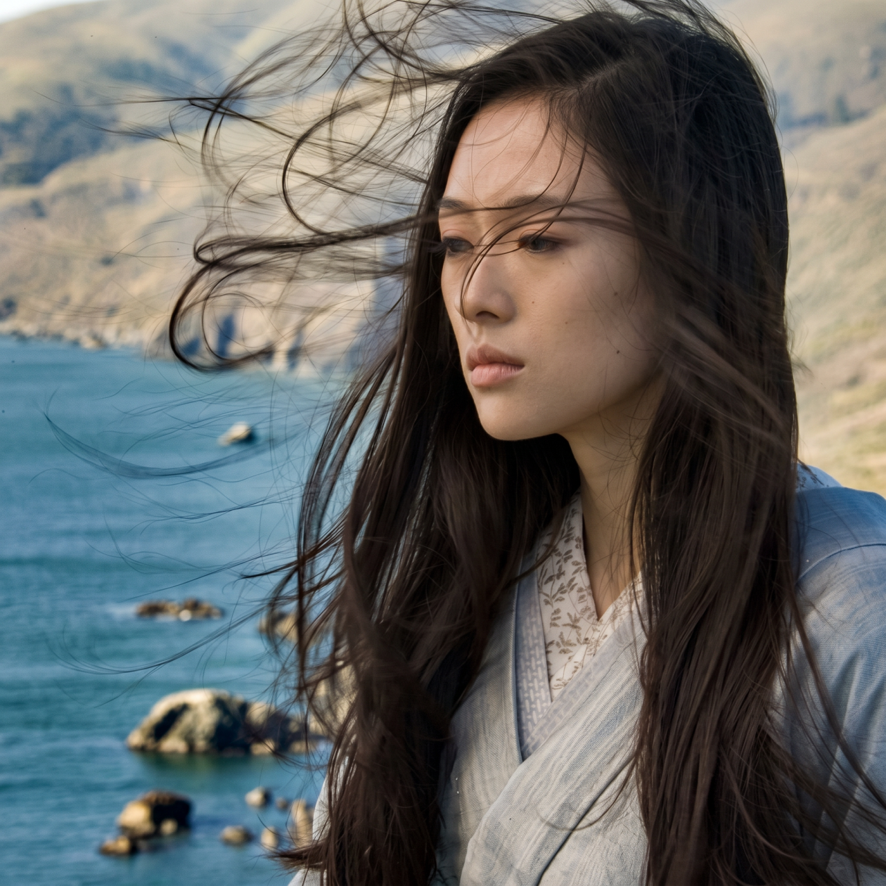 Young woman with windswept long dark hair gazing to the side, wearing a traditional robe, rocky coastline and hills in the background.