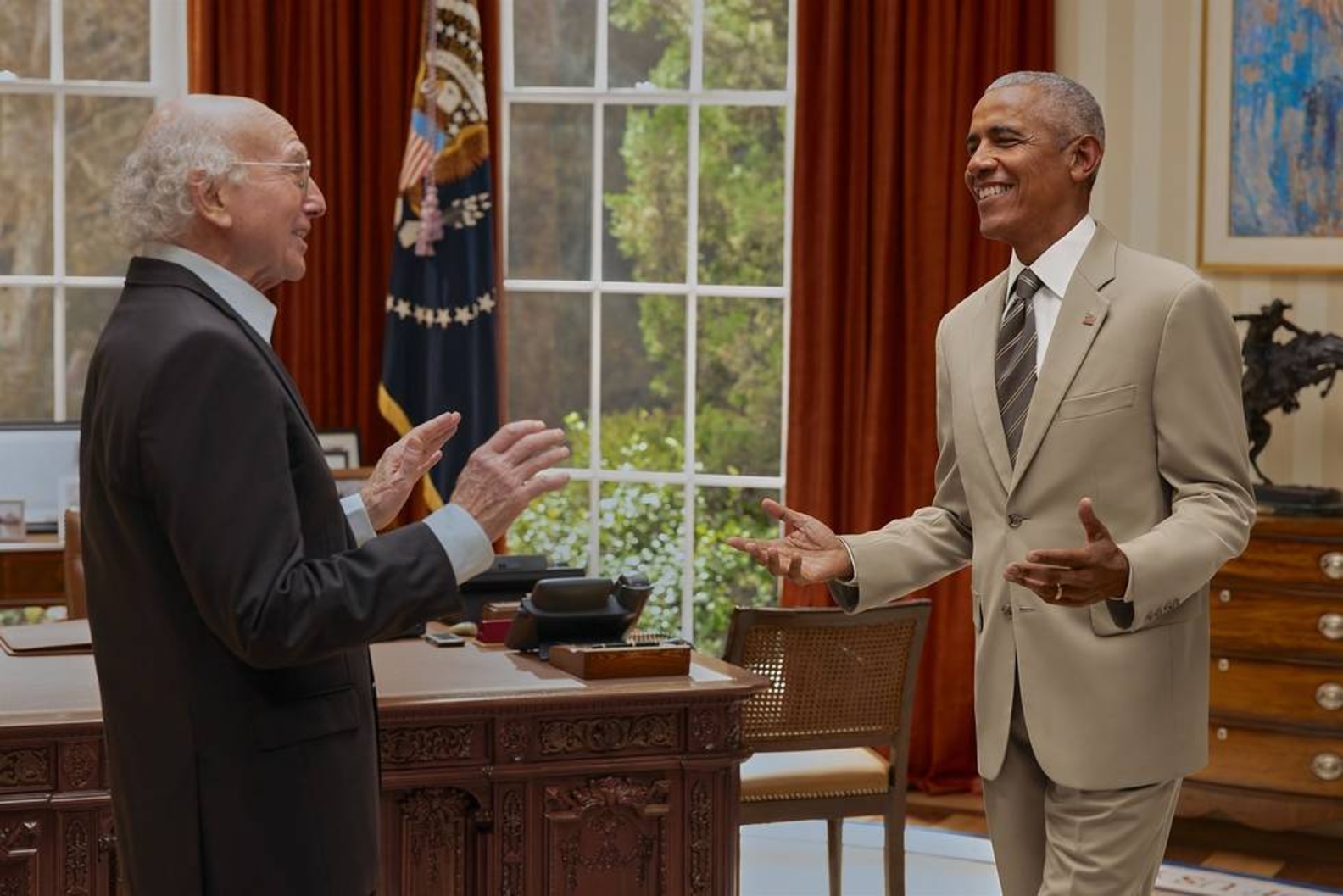 Two men in suits stand across from each other in the Oval Office, both gesturing with their hands mid-conversation, smiling.