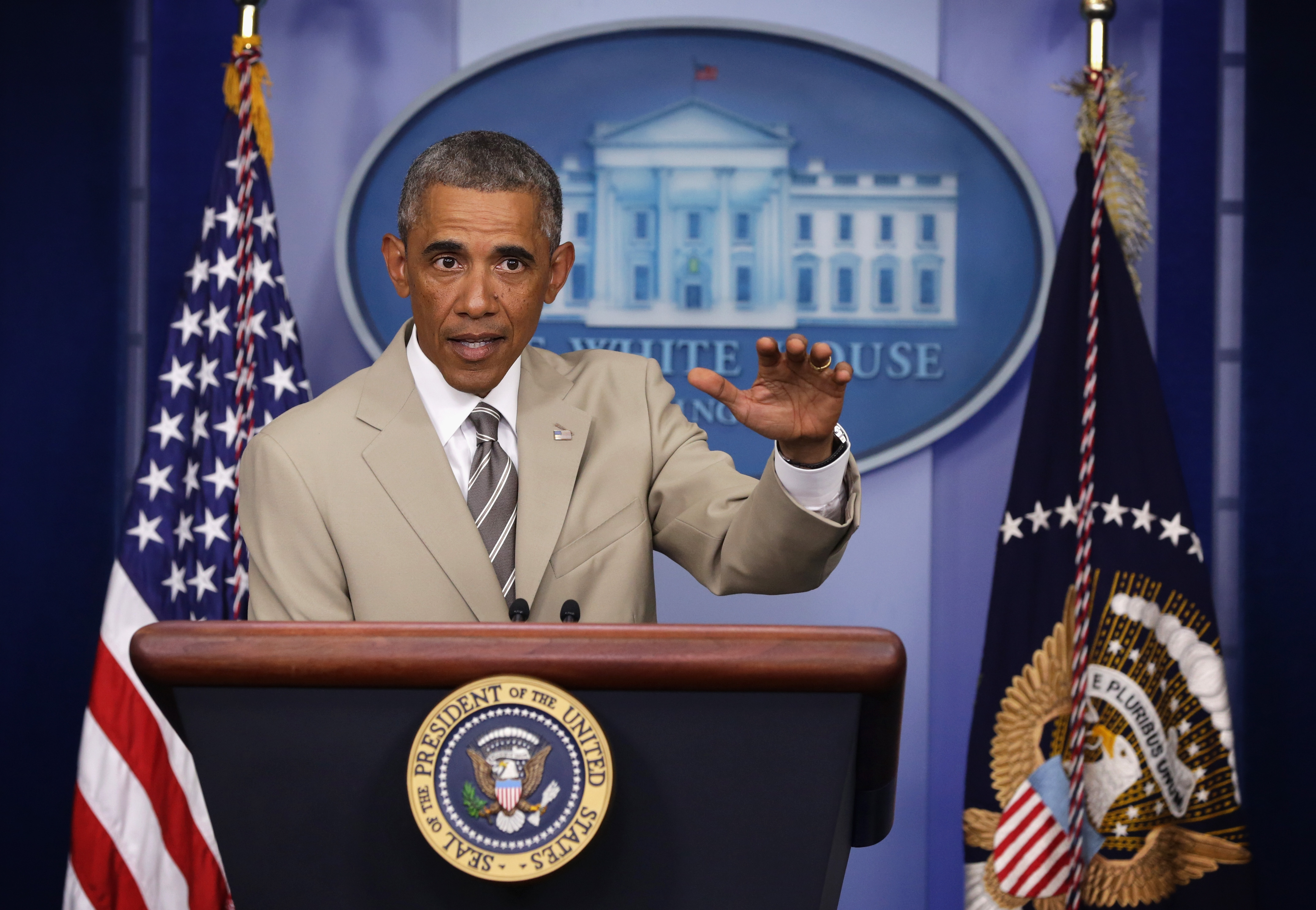 President Obama in a tan suit at the White House briefing room podium, gesturing with one hand, with the Presidential Seal and U.S. flags visible.