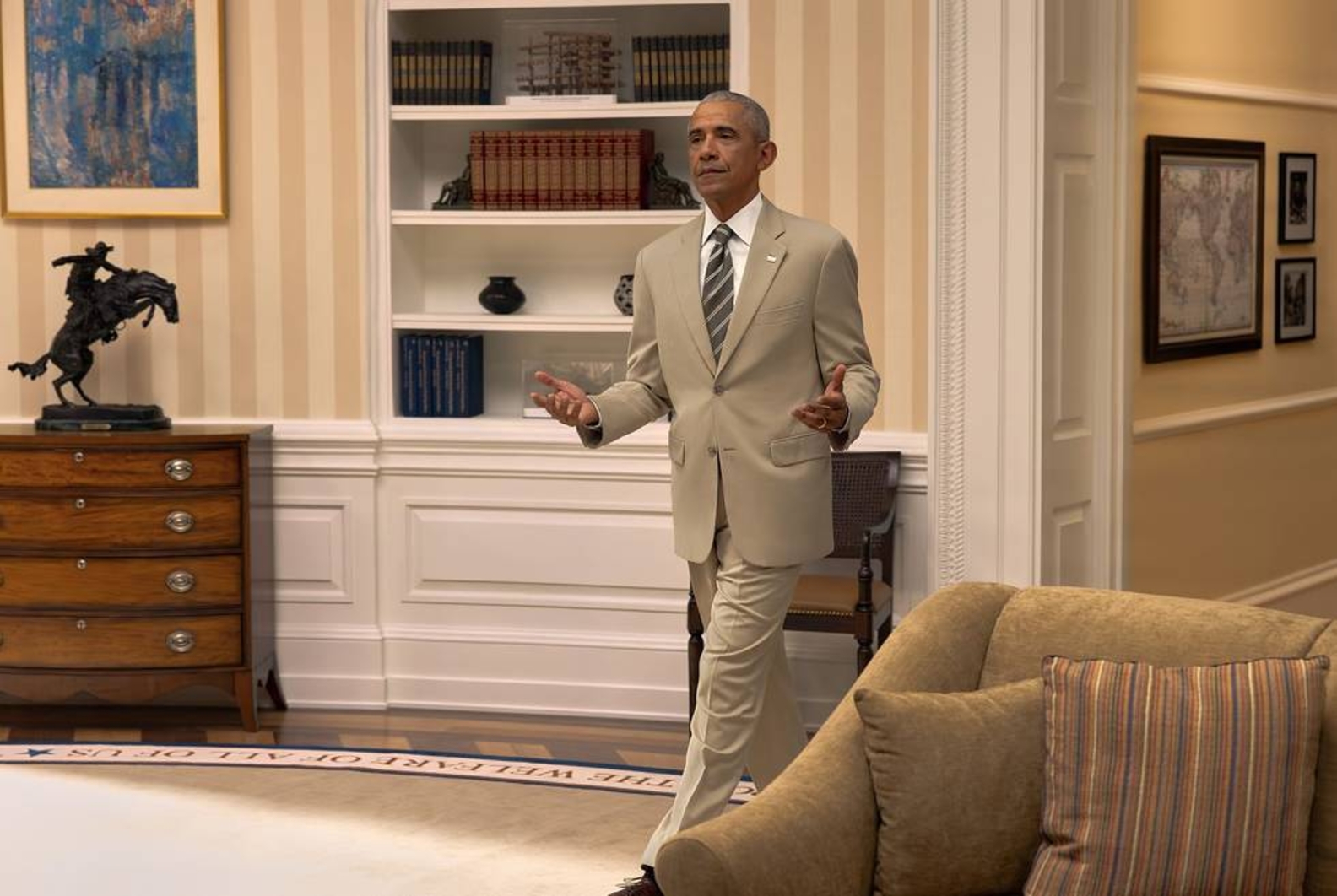 A man in a tan suit gestures with both hands open in a room with a bookcase, a bronze horse sculpture, and a patterned rug border.