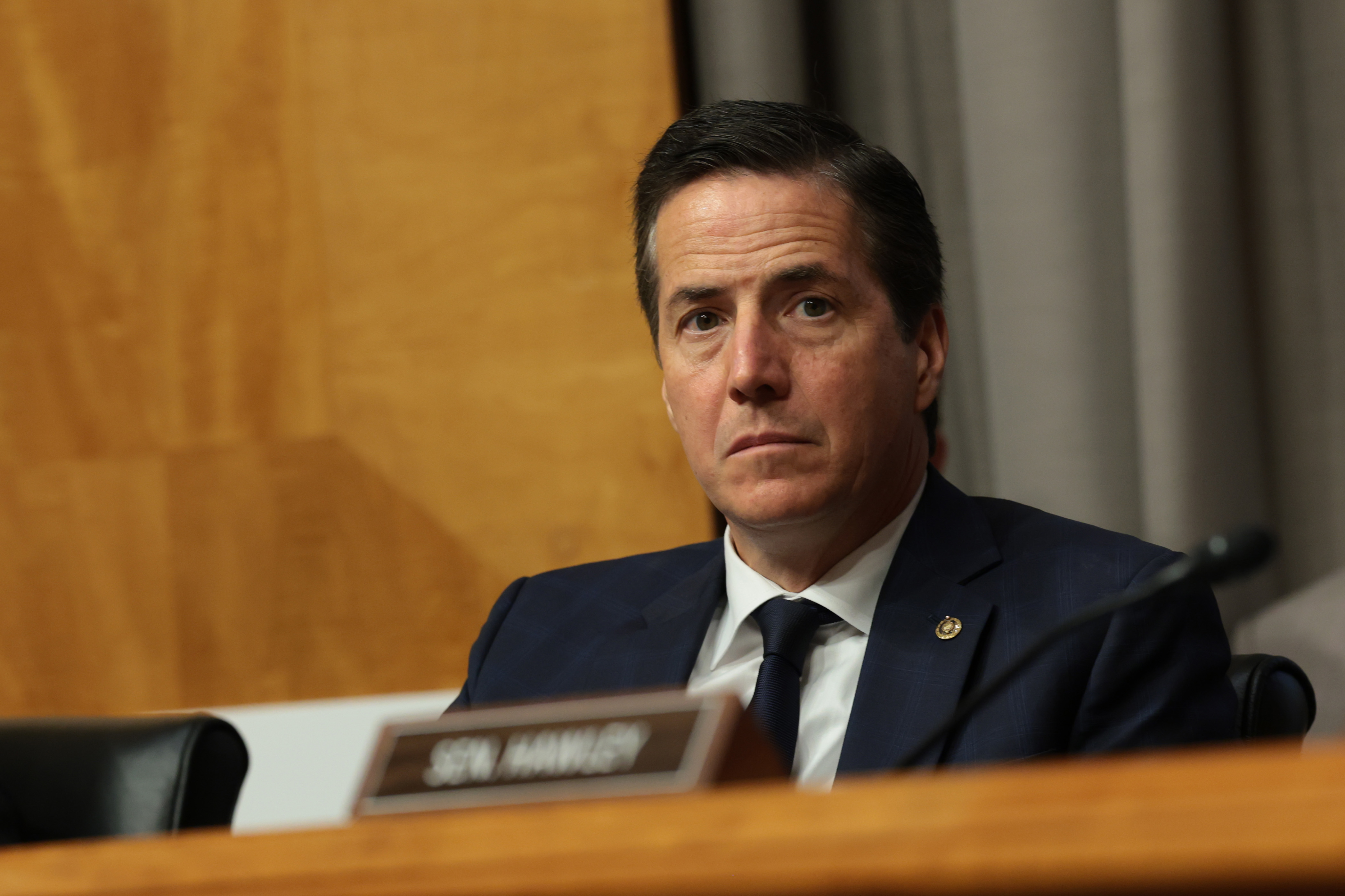 Man in a navy suit and dark tie seated at a Senate committee dais, a nameplate partially visible in the foreground