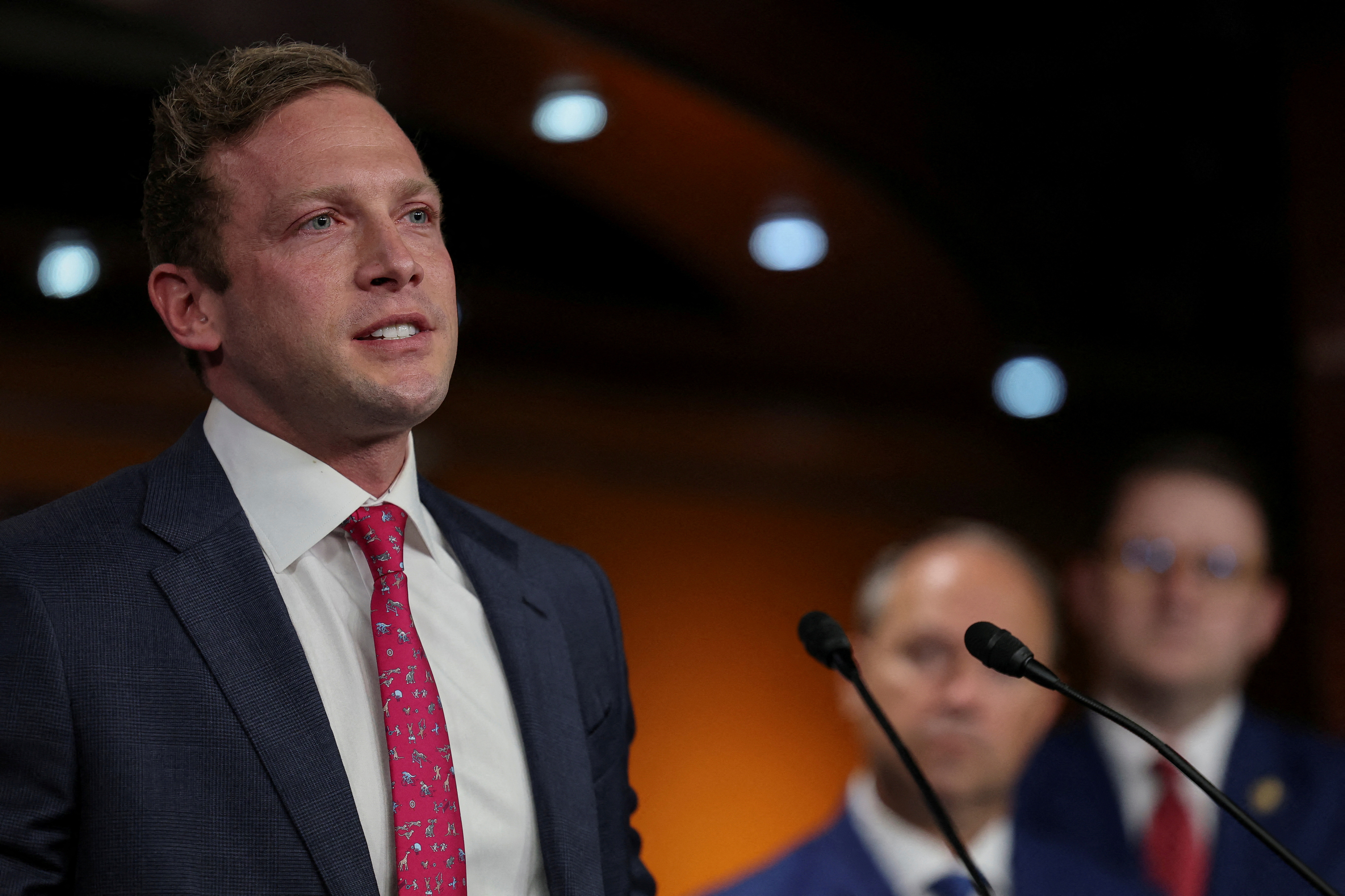 Man in a navy suit and red tie speaking at a podium with microphones, other suited men blurred in background