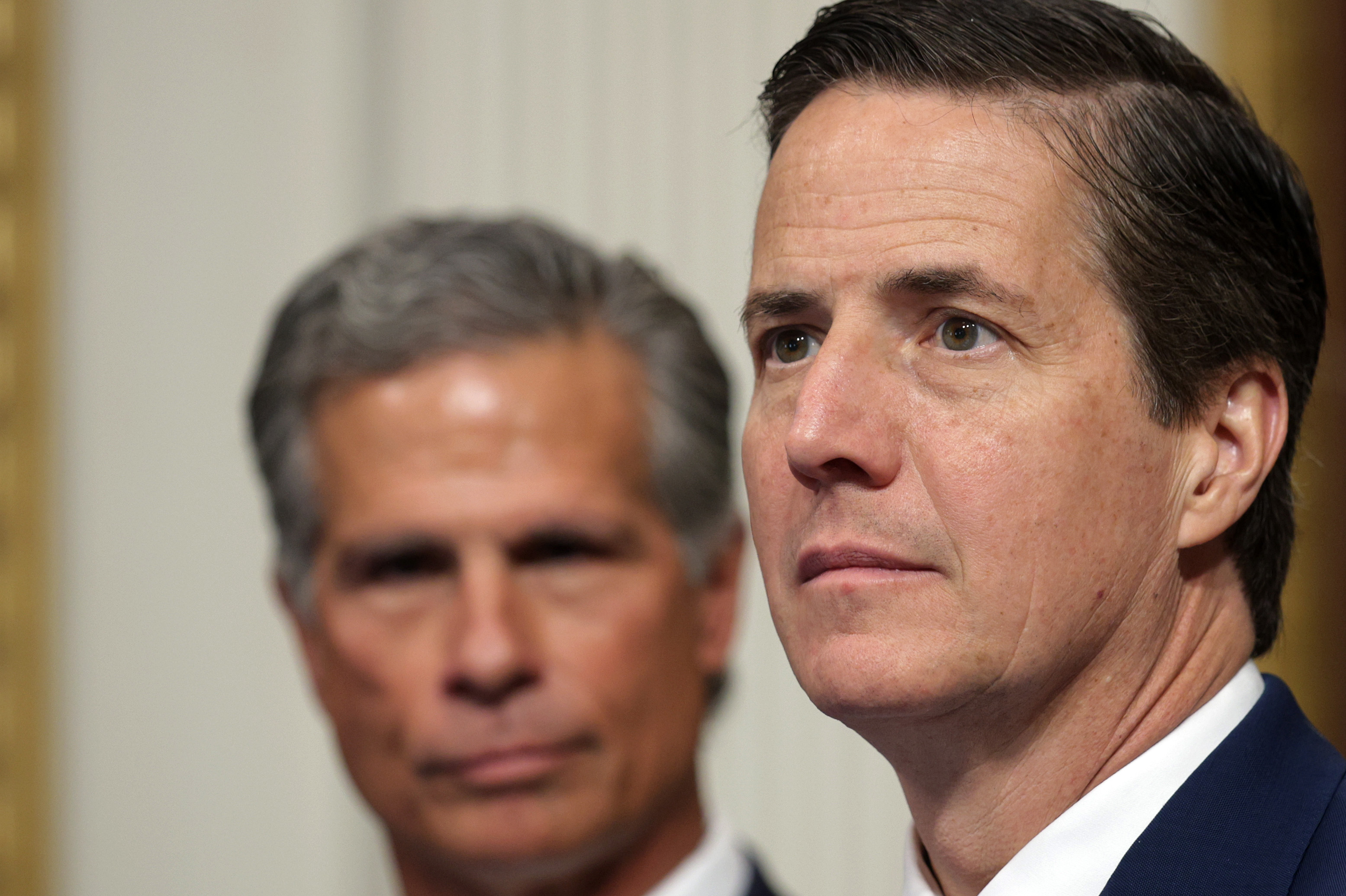 Two men in dark suits at a formal government roundtable, the man on the right in sharp focus looking forward