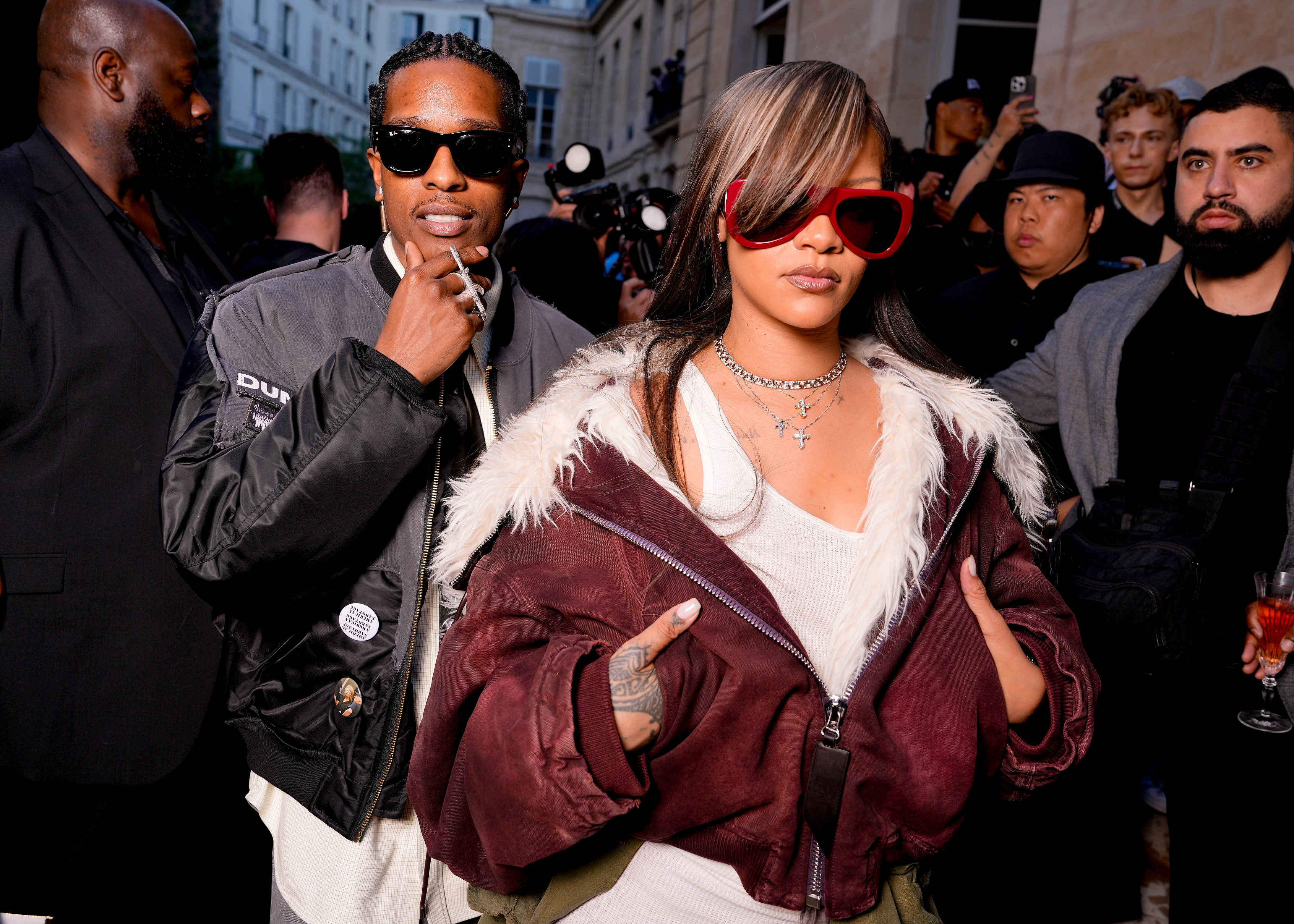 A$AP Rocky in a gray bomber jacket and sunglasses stands beside a woman in a burgundy fur-trimmed jacket and red sunglasses at a Paris Fashion Week event.