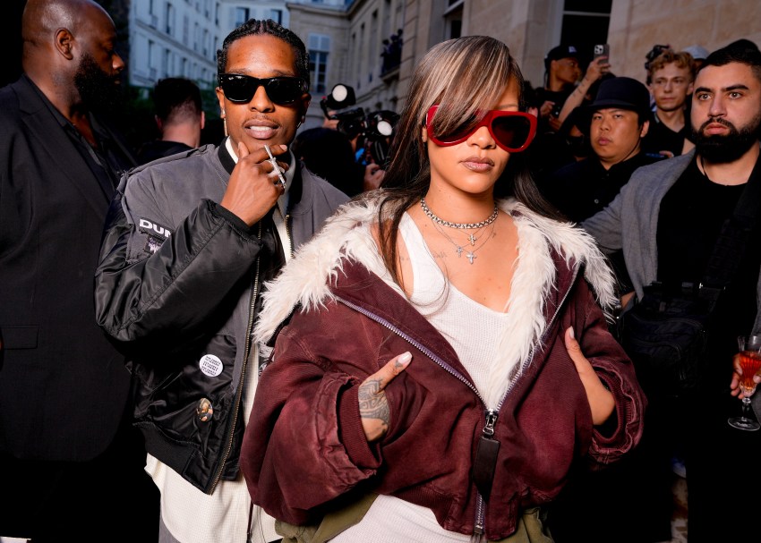 A$AP Rocky in a gray bomber jacket and sunglasses stands beside a woman in a burgundy fur-trimmed jacket and red sunglasses at a Paris Fashion Week event.
