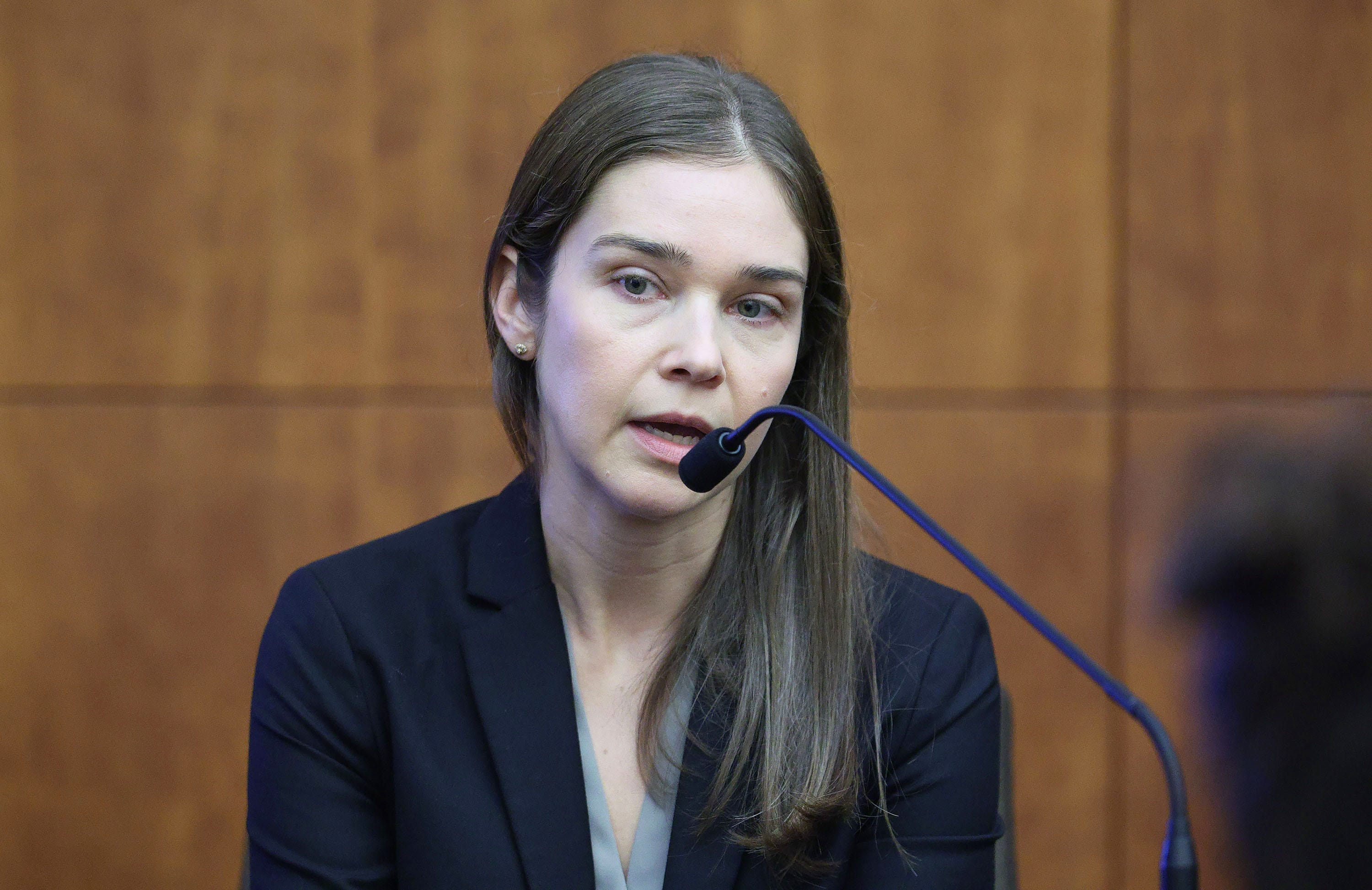 Woman with long brown hair speaking into a microphone at a witness stand in a courtroom, wearing a dark blazer.