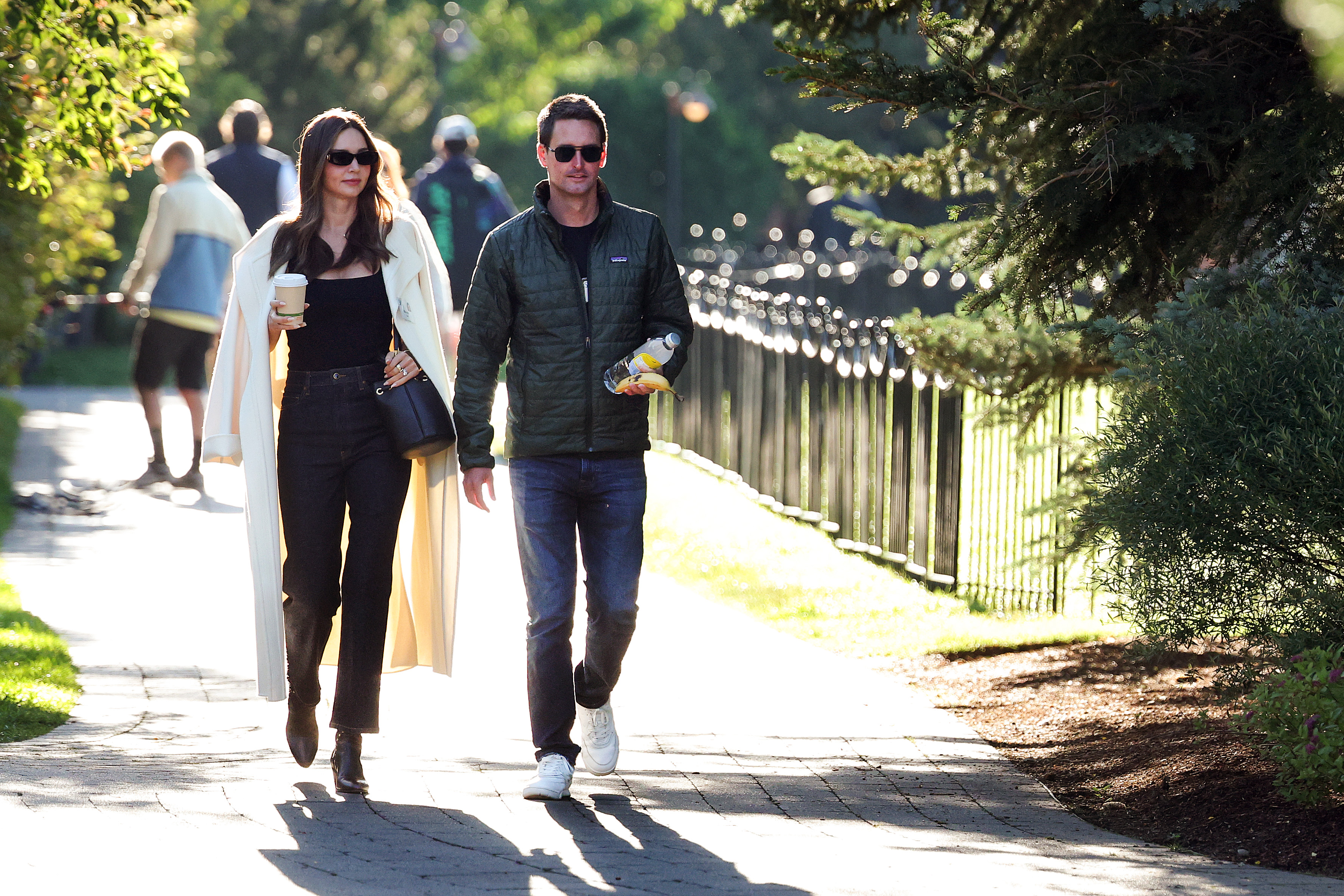 Miranda Kerr and Evan Spiegel walking together on a tree-lined path in Sun Valley, Idaho, both wearing sunglasses.