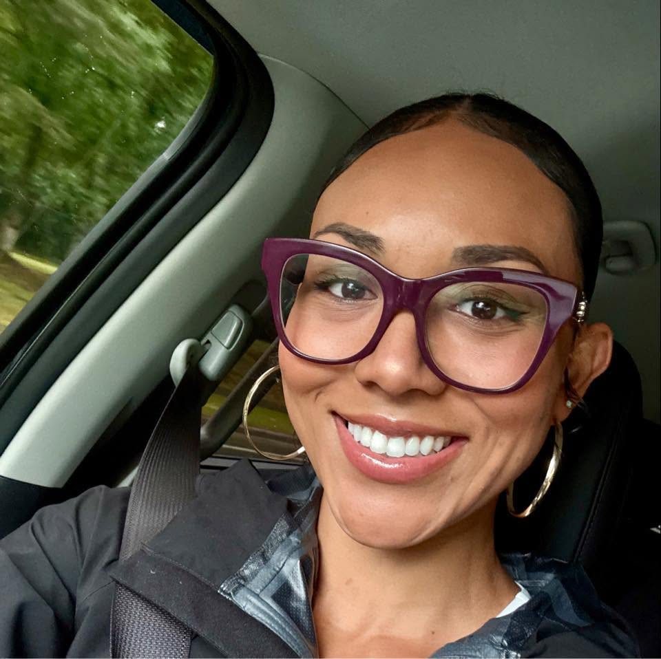 Woman smiling in a car selfie, wearing purple cat-eye glasses and gold hoop earrings, with greenery visible through the window.