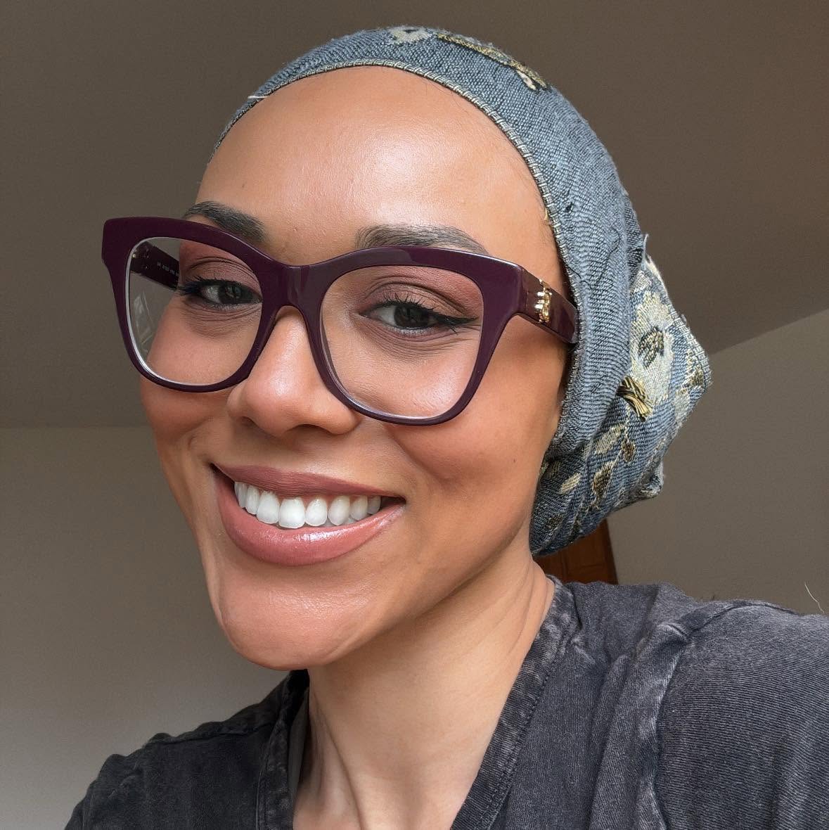 Woman smiling in an indoor selfie, wearing purple glasses and a blue denim patterned head wrap, with a dark gray shirt.