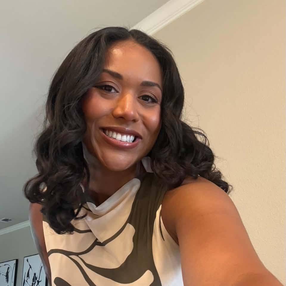 Woman with long wavy dark hair smiling over her shoulder indoors, wearing a sleeveless olive and cream abstract-print top.