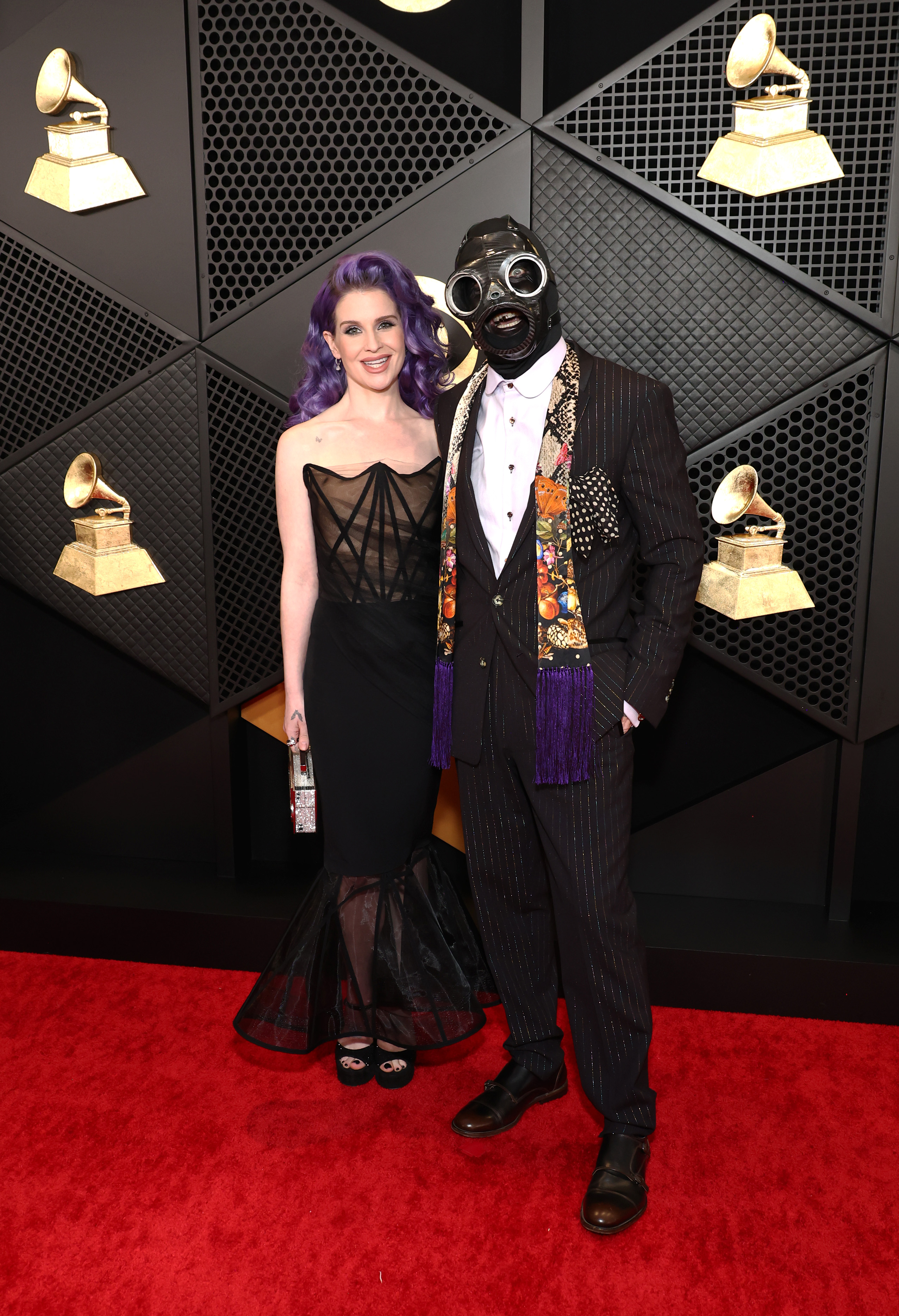 Kelly Osbourne in a black sheer gown with purple hair stands beside Sid Wilson of Slipknot, who wears a pinstripe suit and black gas mask, on the Grammy Awards red carpet.