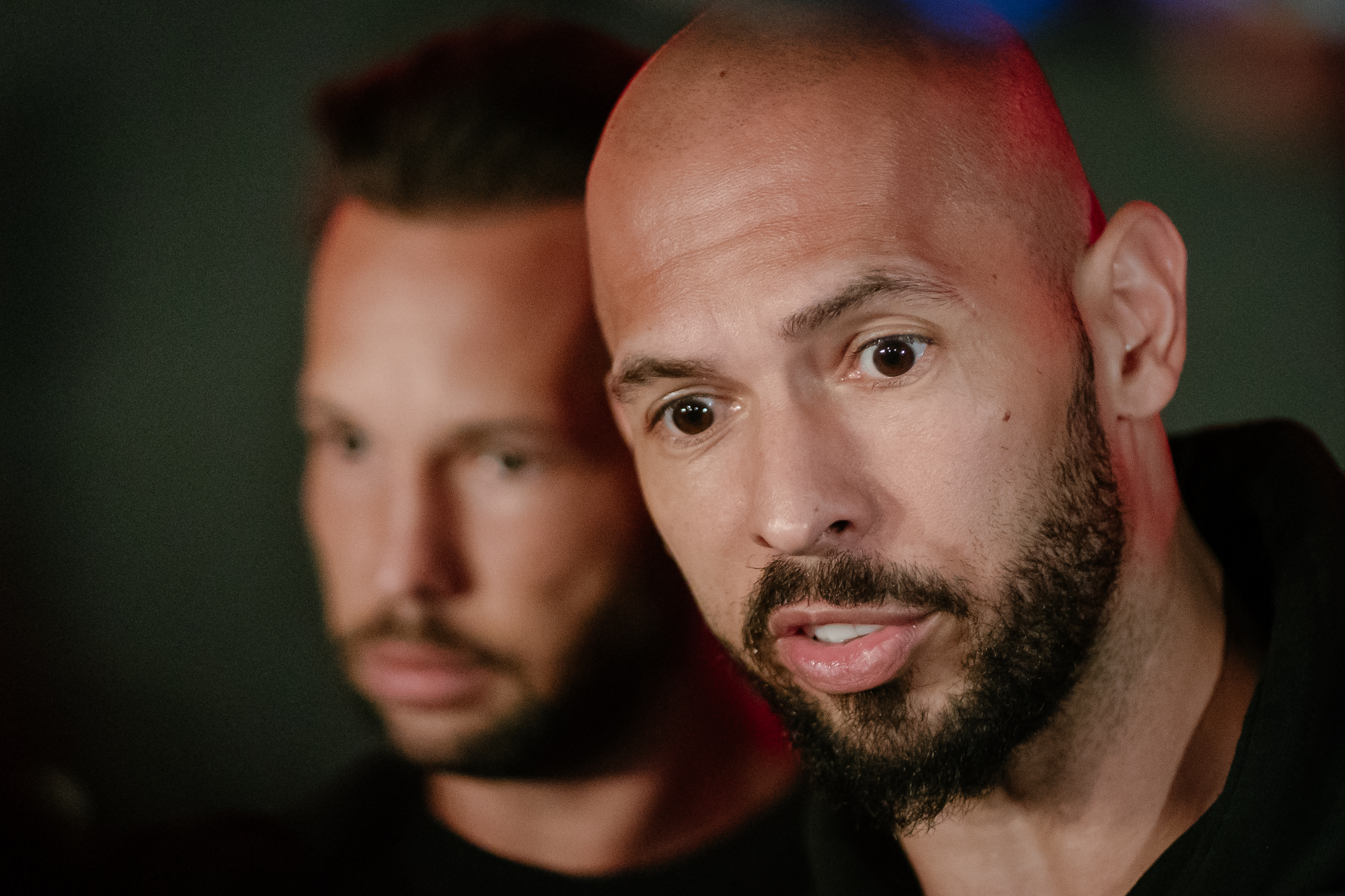 Close-up of a bald man with a dark beard speaking, with another man with short dark hair blurred in the background.