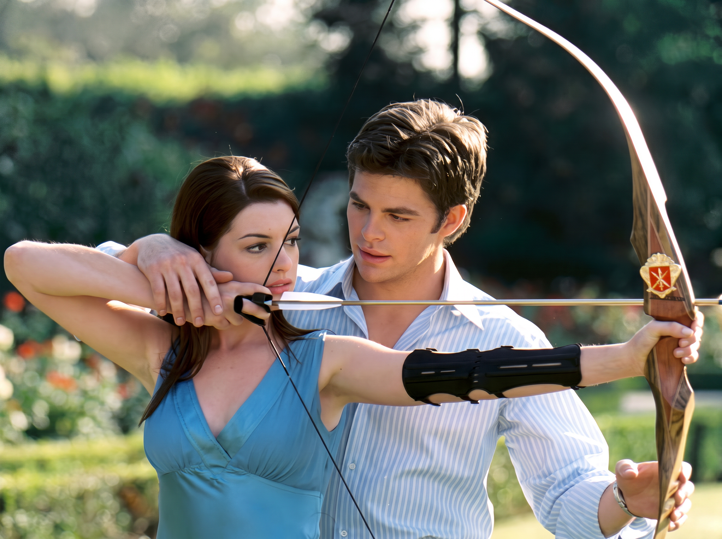 A young man stands behind a woman in a blue dress, helping her aim a bow and arrow outdoors in a garden.
