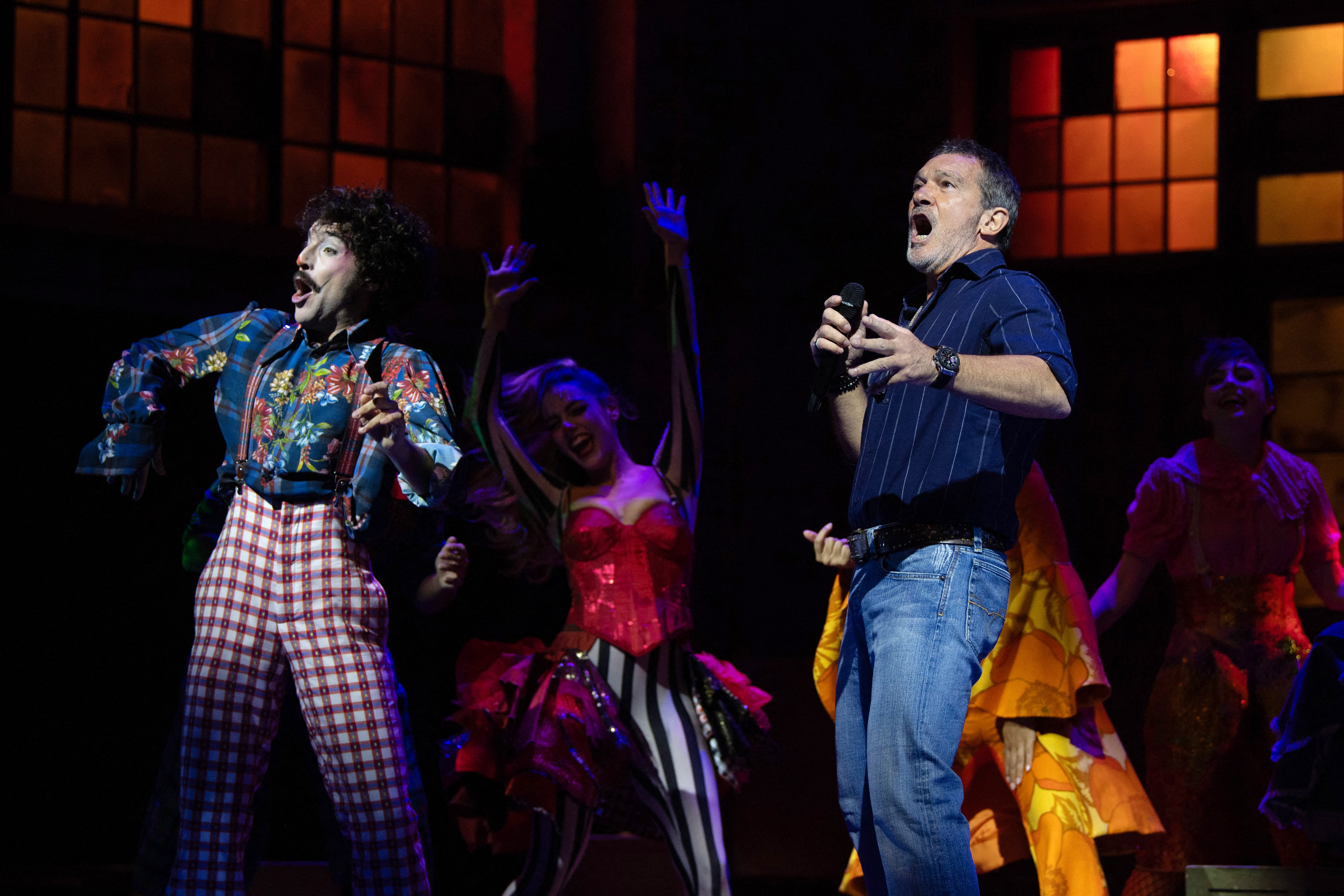 Antonio Banderas singing into a microphone on stage beside performers in vibrant costumes during a theatrical rehearsal.