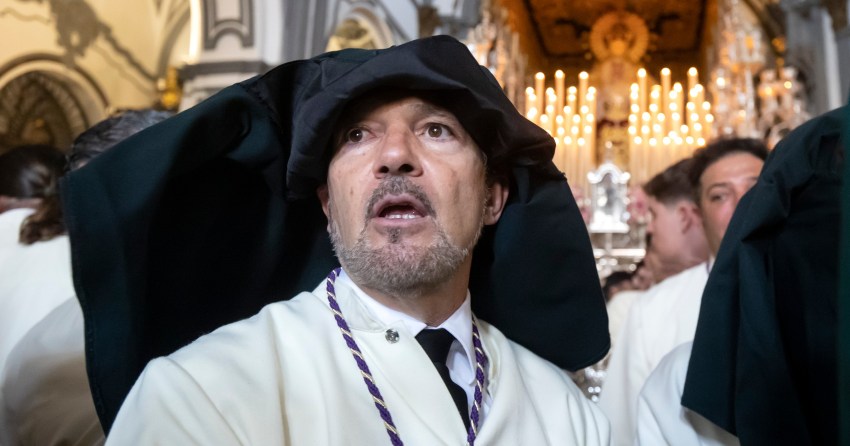 Antonio Banderas in white robes and a black hood looks upward inside an ornate church during a Holy Week procession in Málaga, Spain.