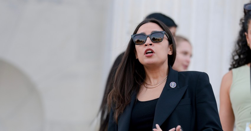 Rep. Alexandria Ocasio-Cortez in a black blazer and sunglasses speaks to reporters on the steps of the U.S. Capitol.
