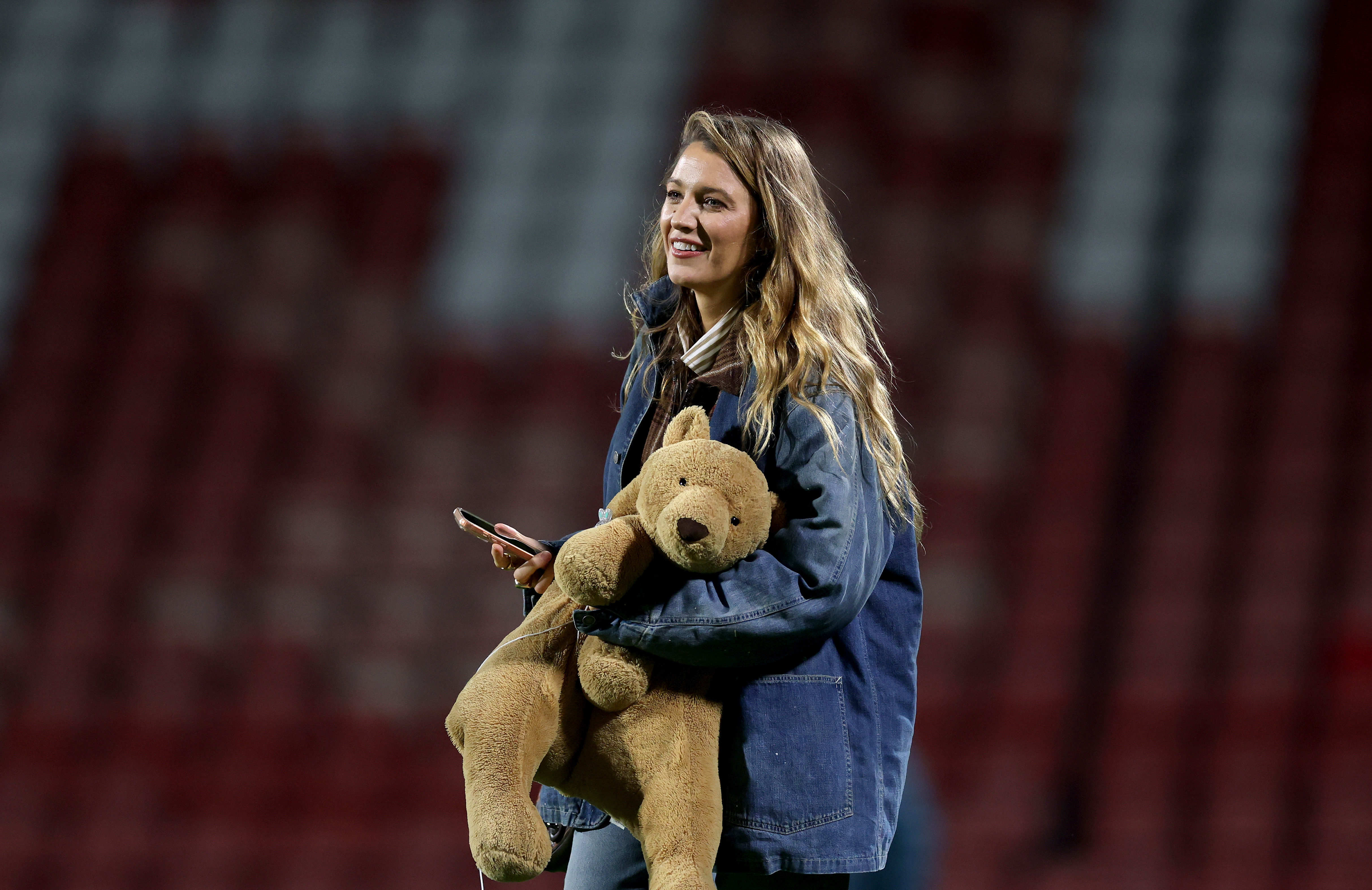 A smiling woman with long wavy hair holds a large stuffed teddy bear and a smartphone, standing in front of empty red stadium seats.