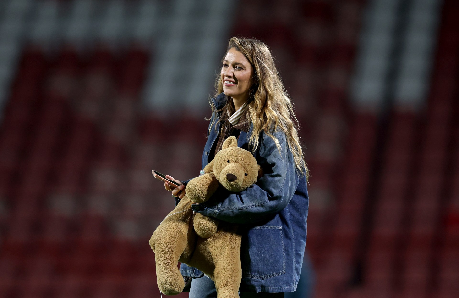 A smiling woman with long wavy hair holds a large stuffed teddy bear and a smartphone, standing in front of empty red stadium seats.
