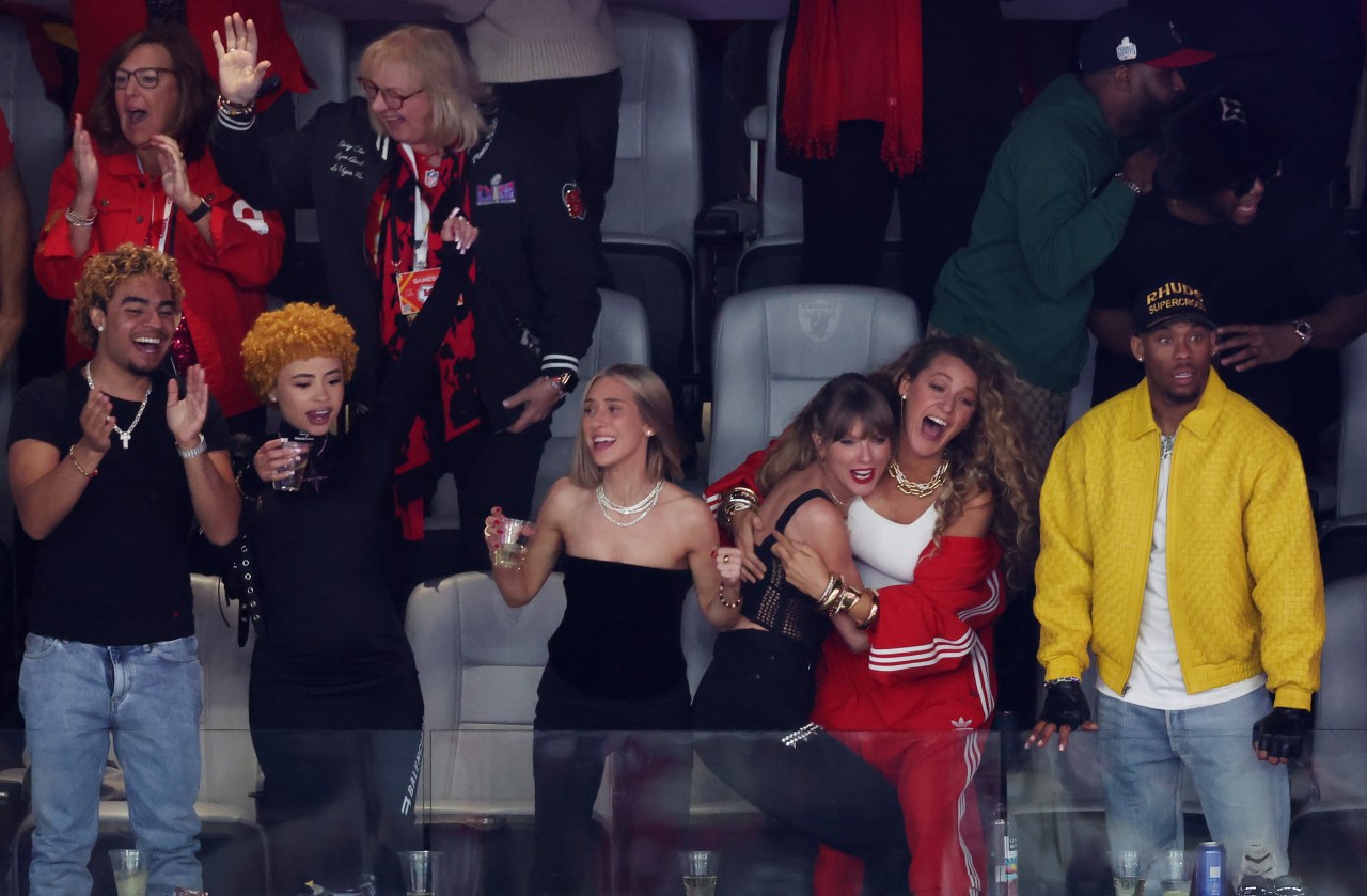 A group of women cheering in stadium seats at Super Bowl LVIII, two of them hugging joyfully, surrounded by friends in a luxury suite.