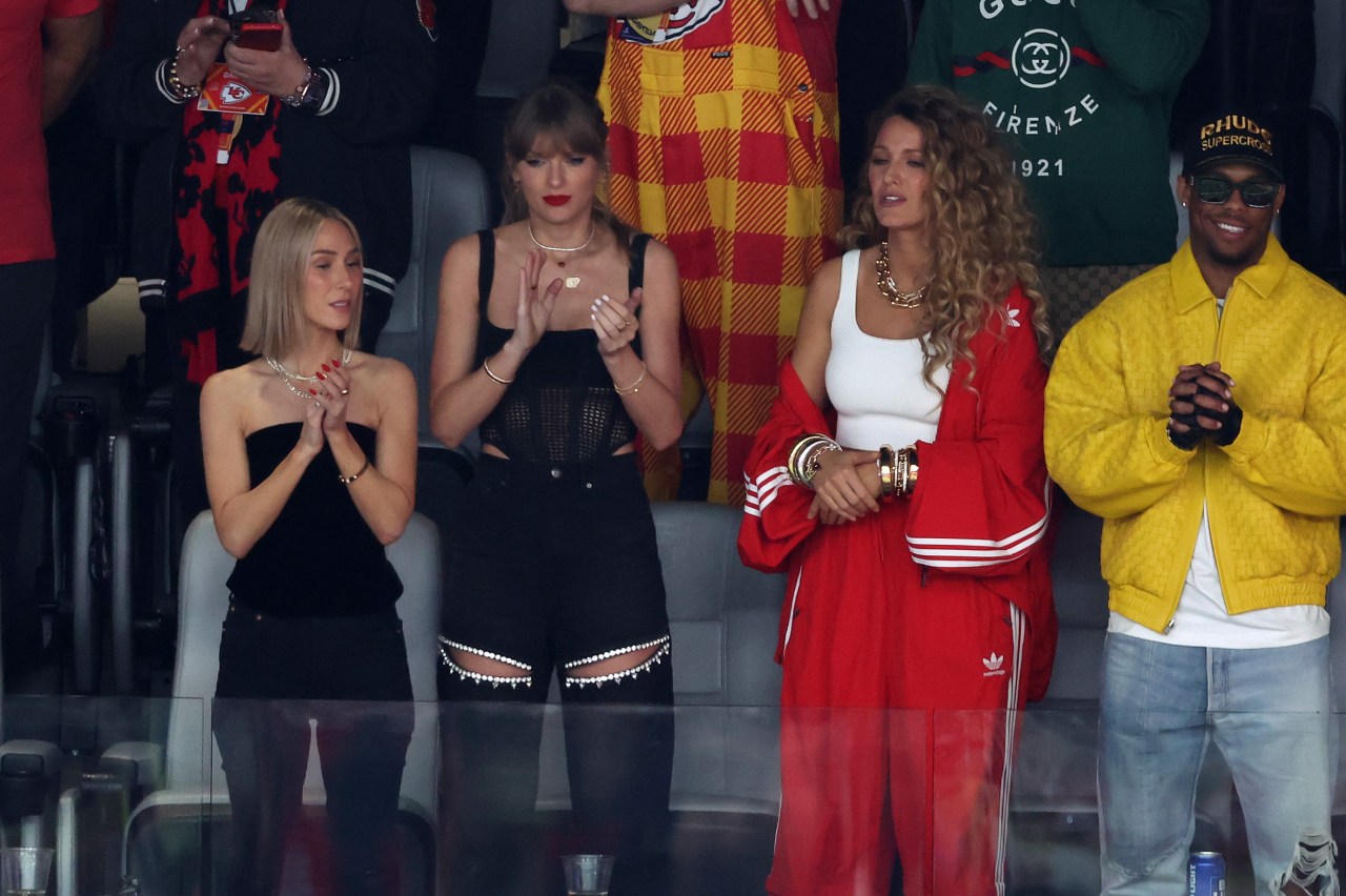 Four people standing and clapping in stadium seats at Super Bowl LVIII; two women in black outfits, one in a red Adidas tracksuit, one man in a yellow jacket.