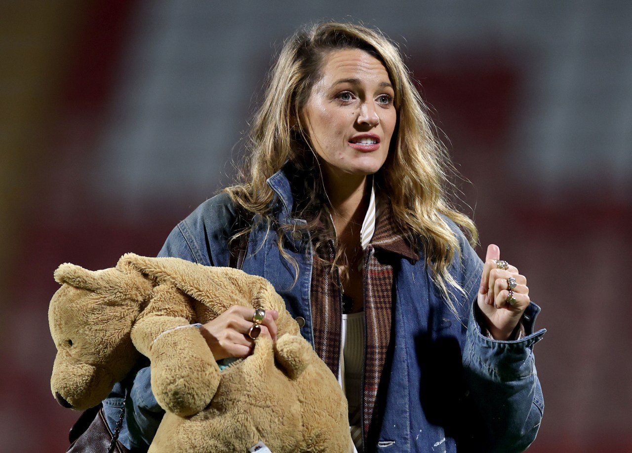 A woman with wavy blonde hair in a denim jacket holds a large stuffed teddy bear on a football pitch, with stadium seating visible in the background.