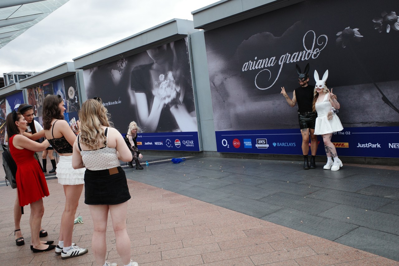 Groups of fans in concert outfits gather outside the O2 Arena; two people wearing bunny masks pose in front of an Ariana Grande billboard while others watch and take photos.