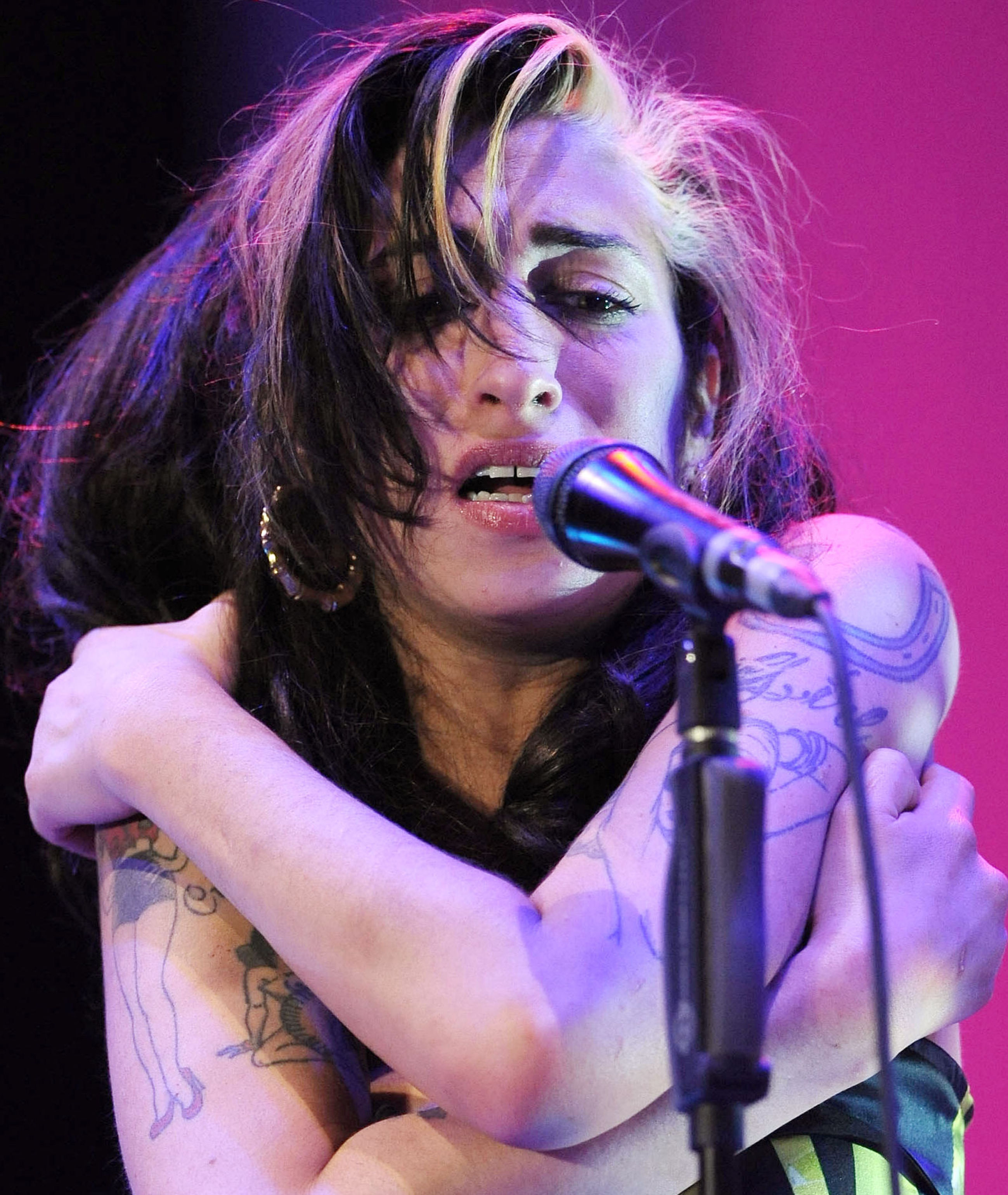 A heavily tattooed woman with disheveled dark hair hugs herself while singing into a microphone on a stage lit in pink and purple.