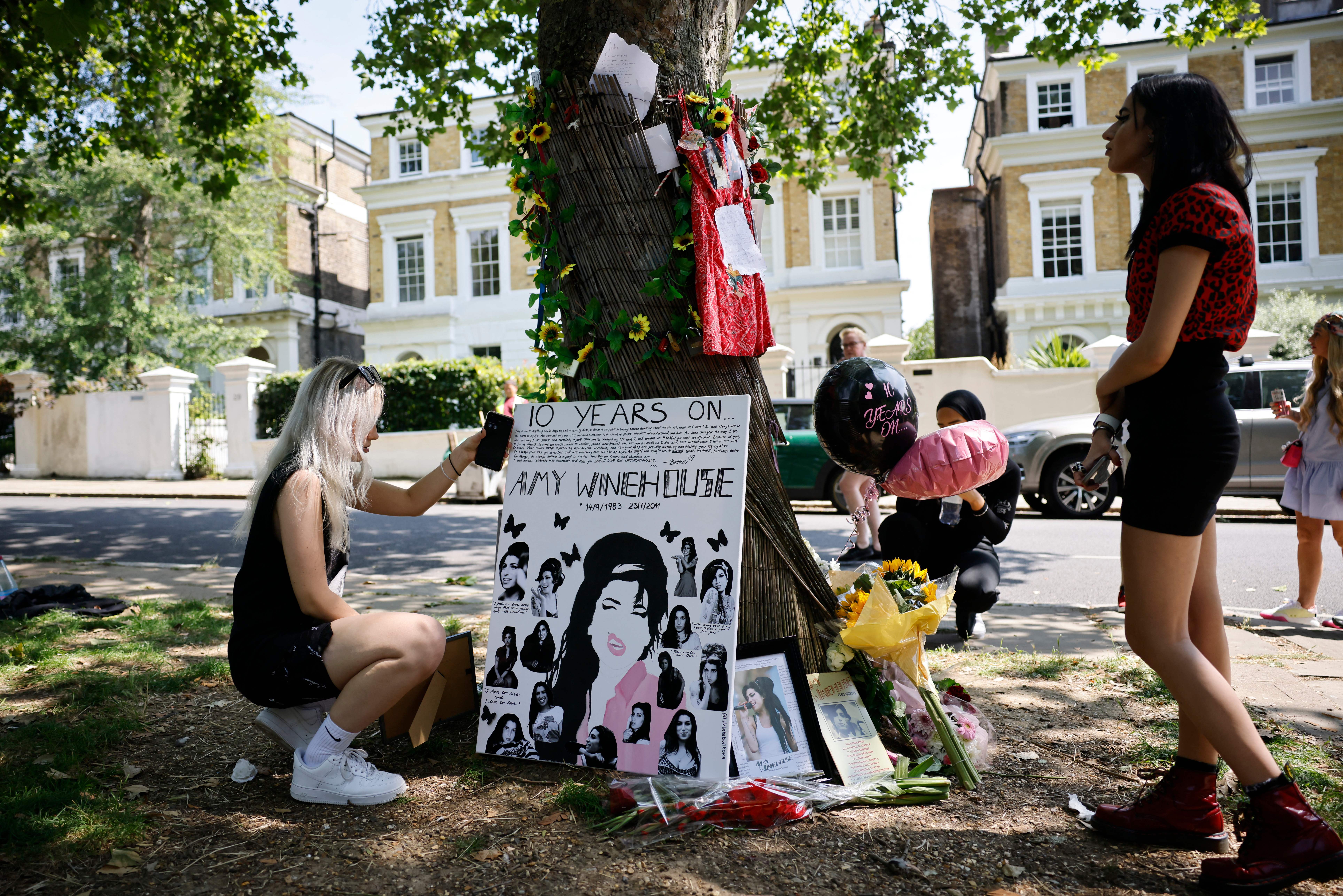 Fans photograph a handmade Amy Winehouse memorial poster at a tree shrine in north London, with flowers and balloons marking the 10th anniversary of her death.