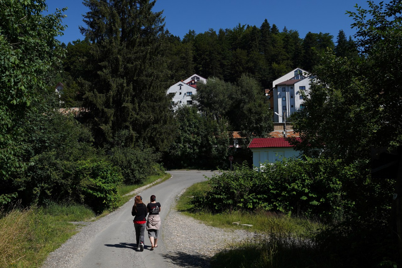 Two women walking away from camera on a narrow gravel road lined with dense green trees, with white buildings visible in the background.