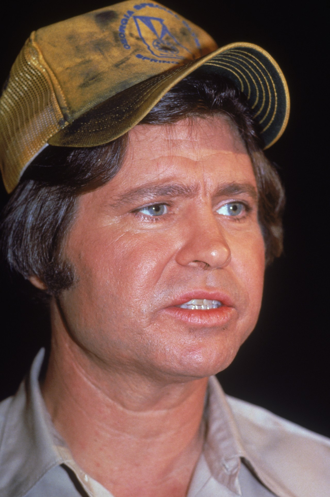 Close-up portrait of Ben Jones wearing a dirty yellow trucker cap and a light button-up shirt, looking slightly off camera, 1983.